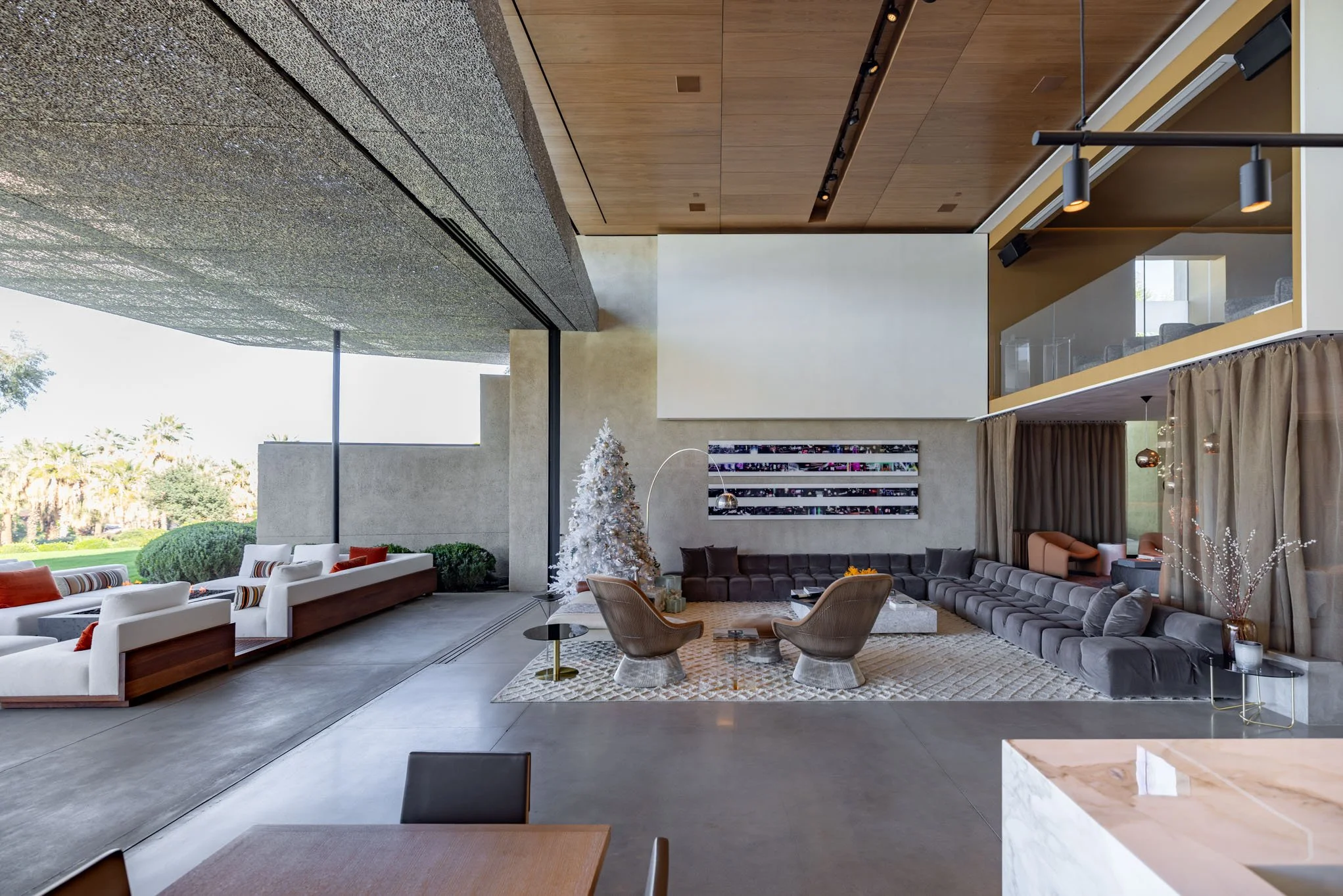 Modern living room with large gray sectional sofa, two beige armchairs, a Christmas tree, and a patterned rug. Large windows and glass wall connect to outdoor greenery.