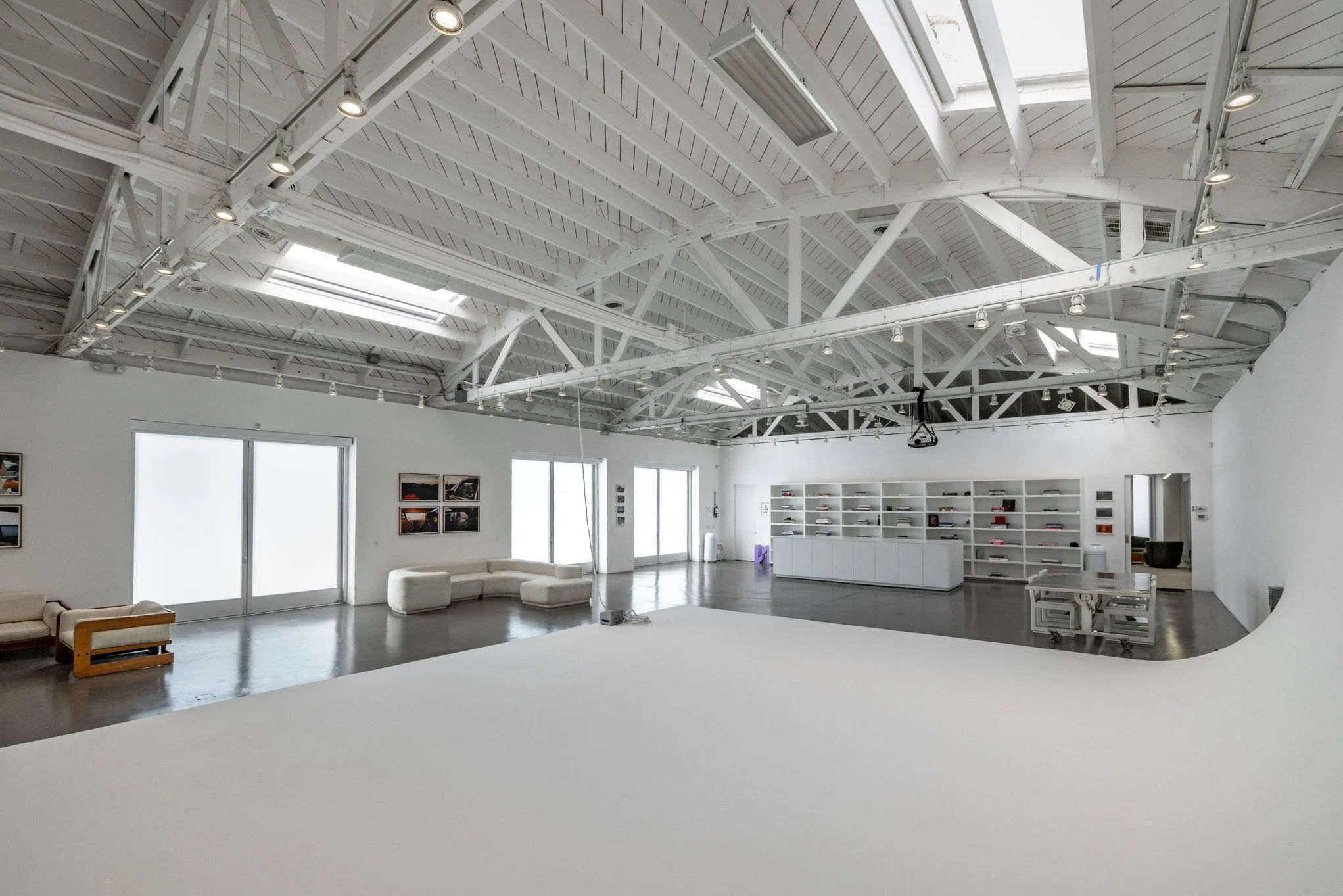 A spacious art gallery with a white painted ceiling featuring exposed beams, skylights, and track lighting. The gallery has large frosted glass doors, framed artwork on the walls, a white curved sofa, and a bookshelf filled with books and art objects