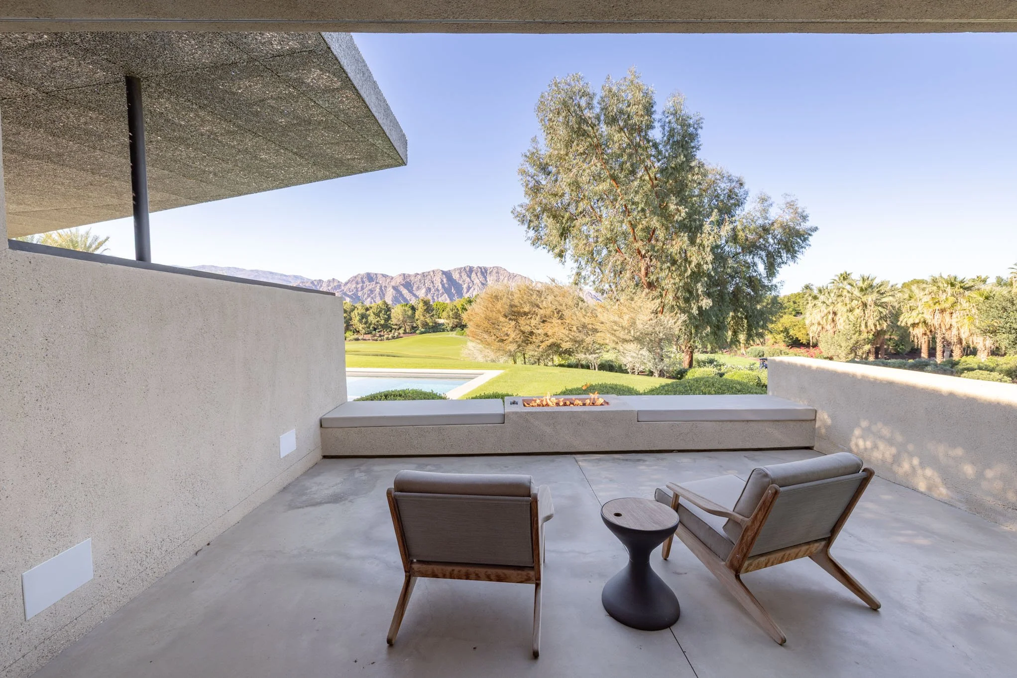 Modern balcony with two armchairs and a small table overlooking a landscape with trees, mountains, and a golf course under a clear blue sky.