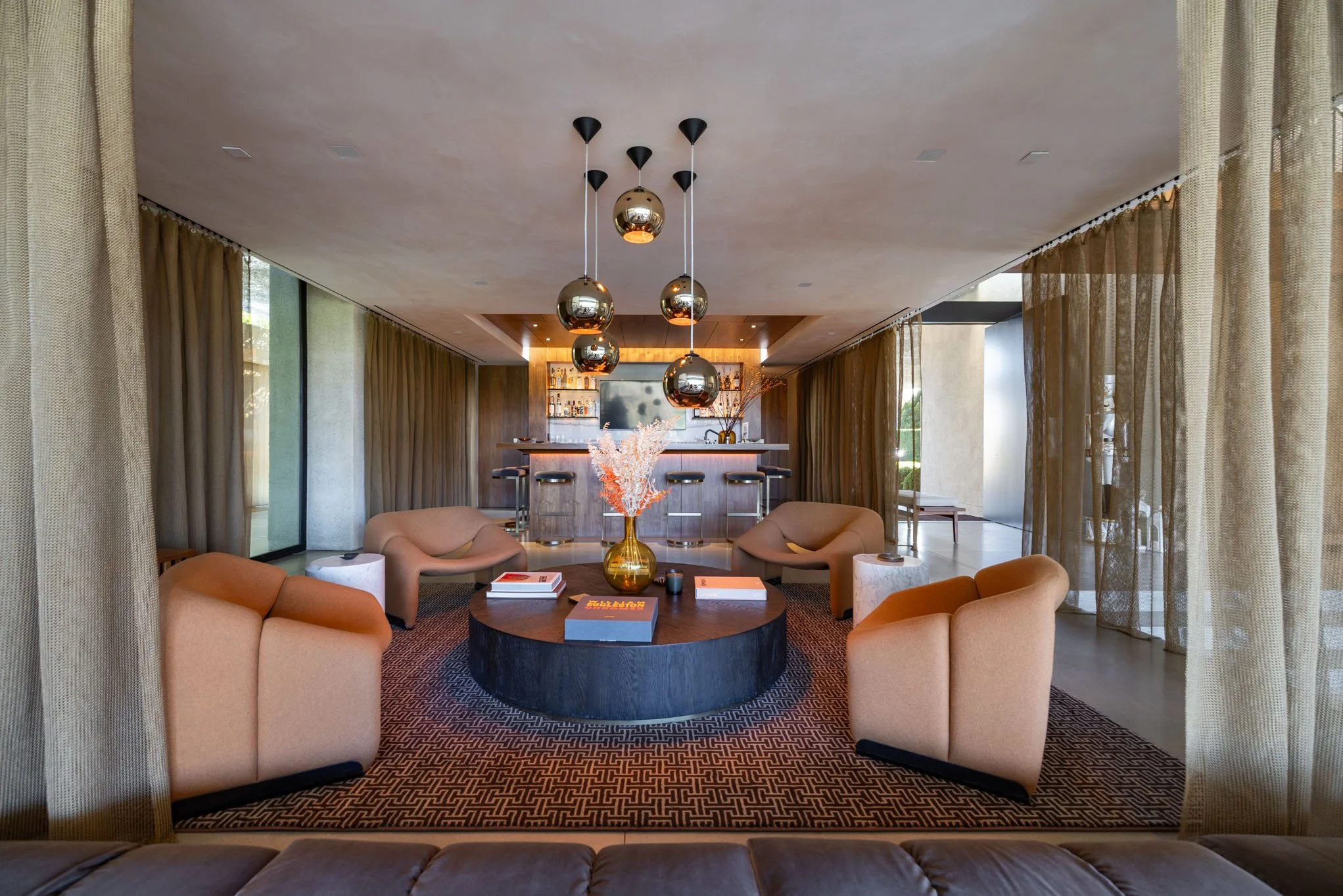 Modern hotel lounge with a central round coffee table, pink armchairs, and a bar in the background, with gold pendant lights hanging from the ceiling.