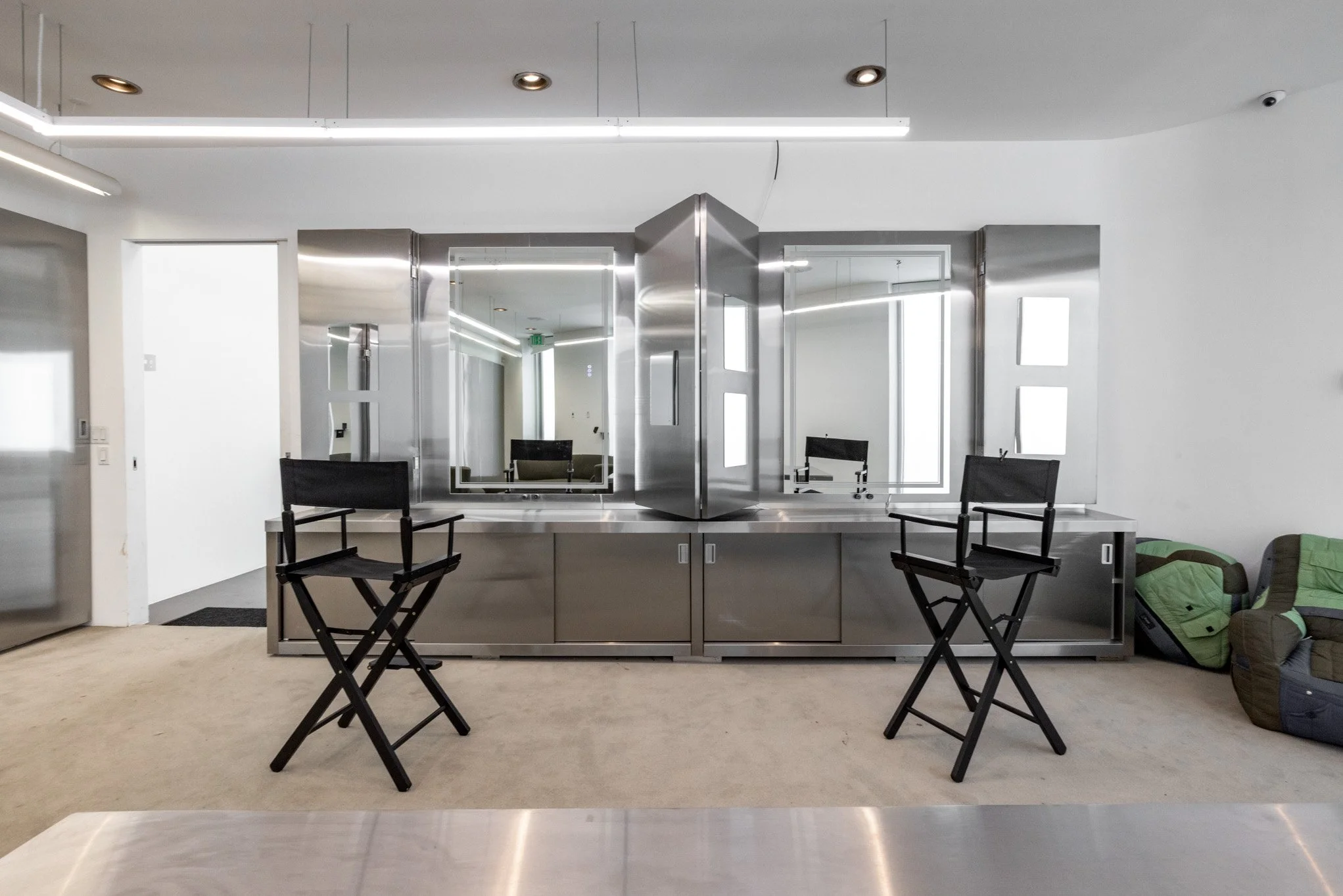 Modern salon station with large mirrors, black director chairs, and stainless steel cabinetry in a bright, minimalist space.