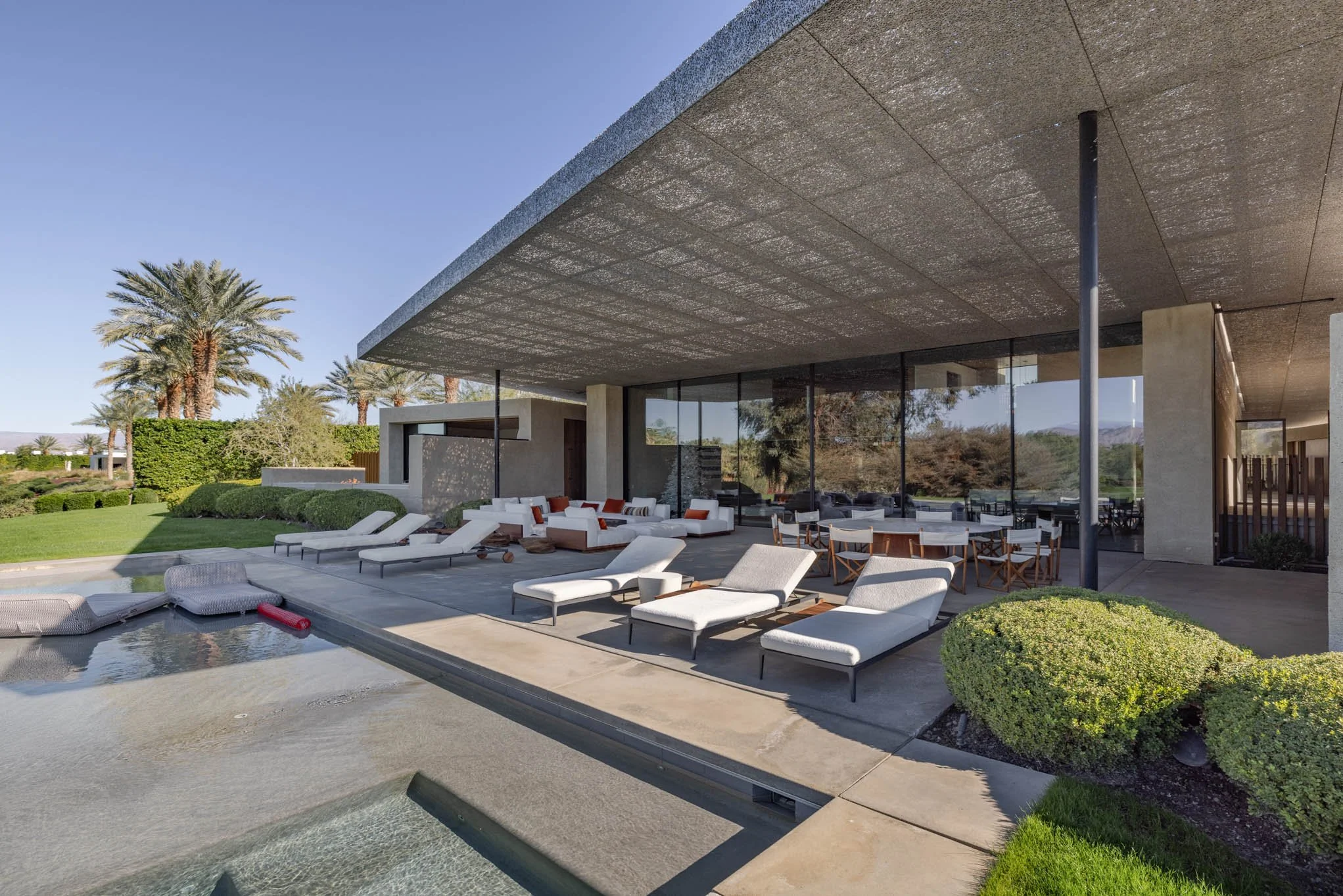 Modern house with a large patio area featuring multiple white lounge chairs, cushioned outdoor seating, a dining table with chairs, and a swimming pool with inflatables. Palm trees and green landscape in the background under a clear blue sky.