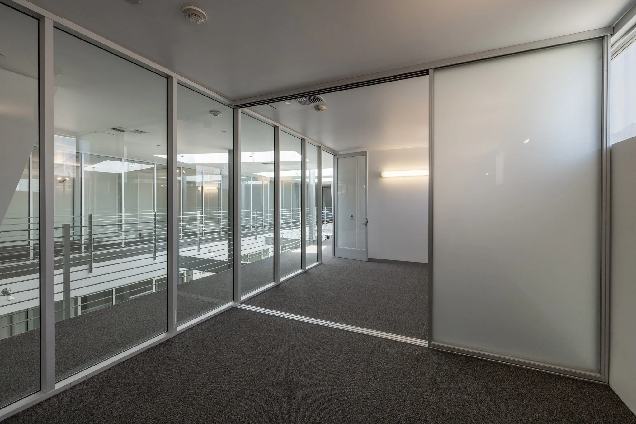 An office space with glass walls, carpeted floor, a door, and fluorescent lighting, overlooking a multi-story building's interior corridor.