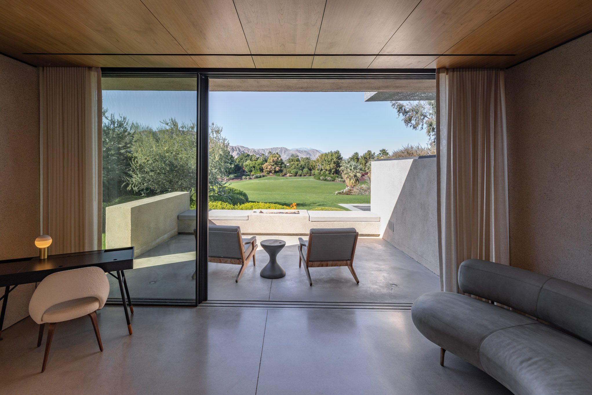 Room with large sliding glass door opening to a view of a green golf course with mountains in the background. Inside, there are two chairs with a small round table between them, a cushioned sofa, a desk with a chair, and curtains pulled to the sides.