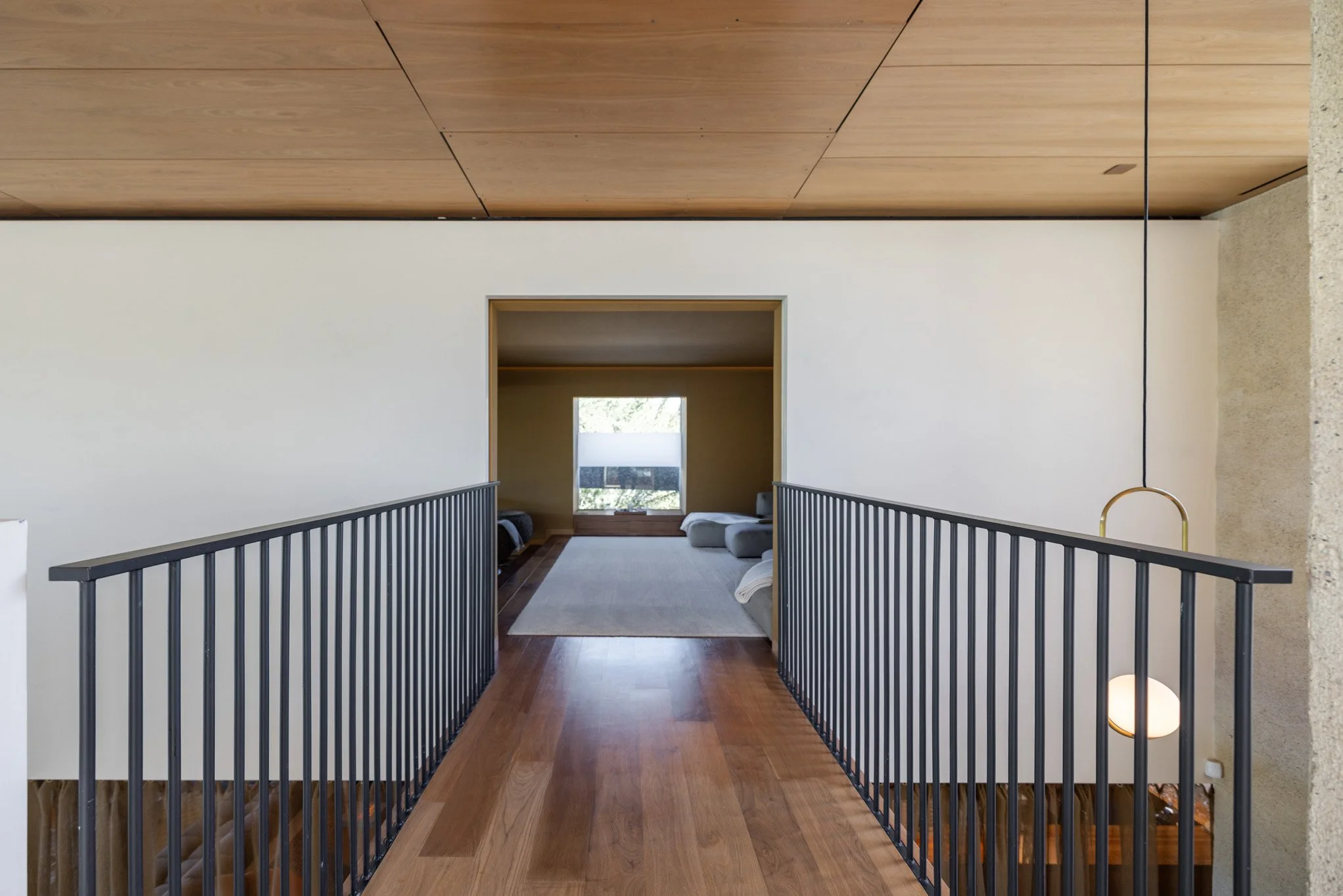 Interior view of a modern house with a second-floor hallway leading to a sitting area with a large window and minimalist furniture.