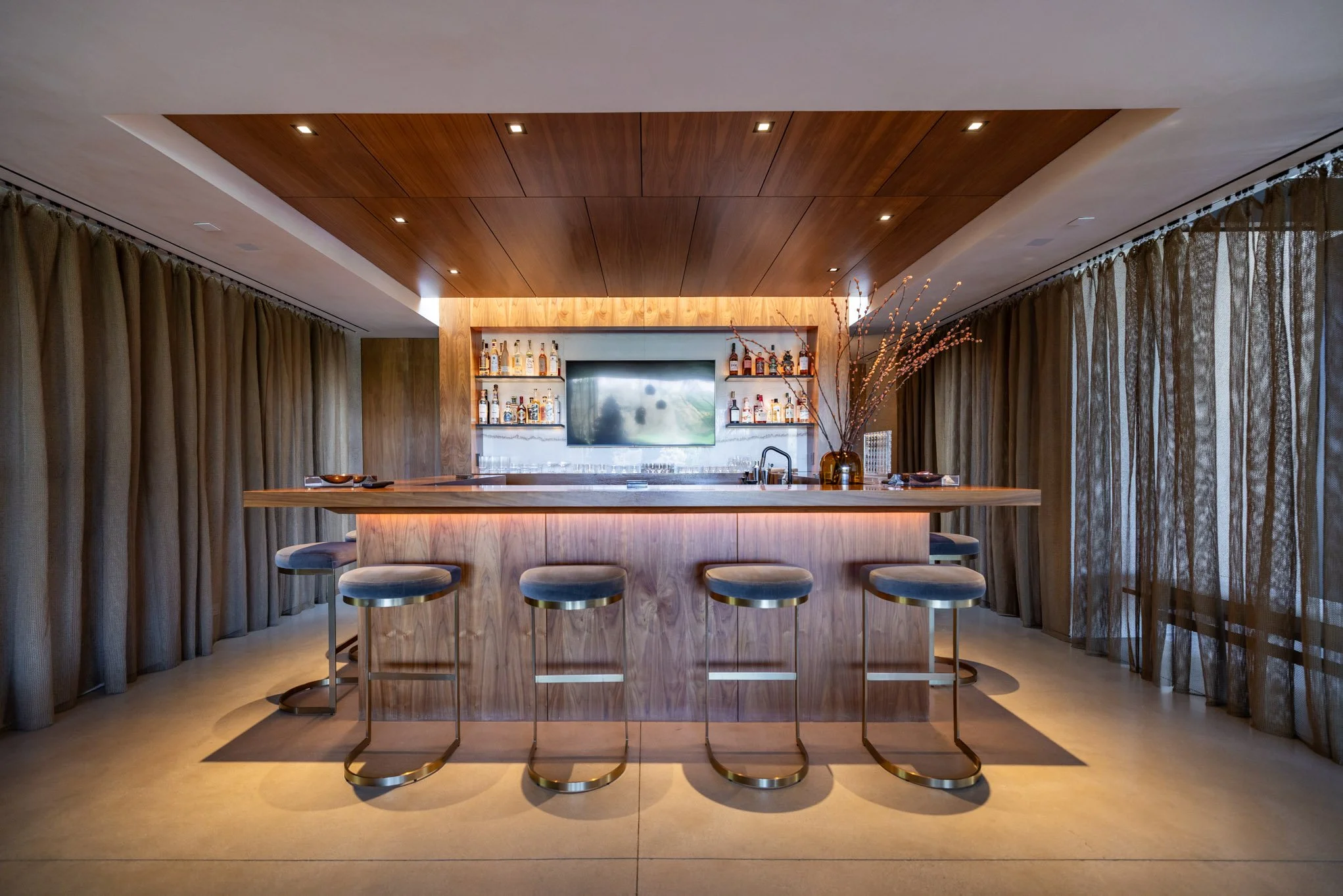 Modern bar with a wooden counter, bar stools, and a wall-mounted TV, featuring curtains on windows and a vase with branches on the counter.