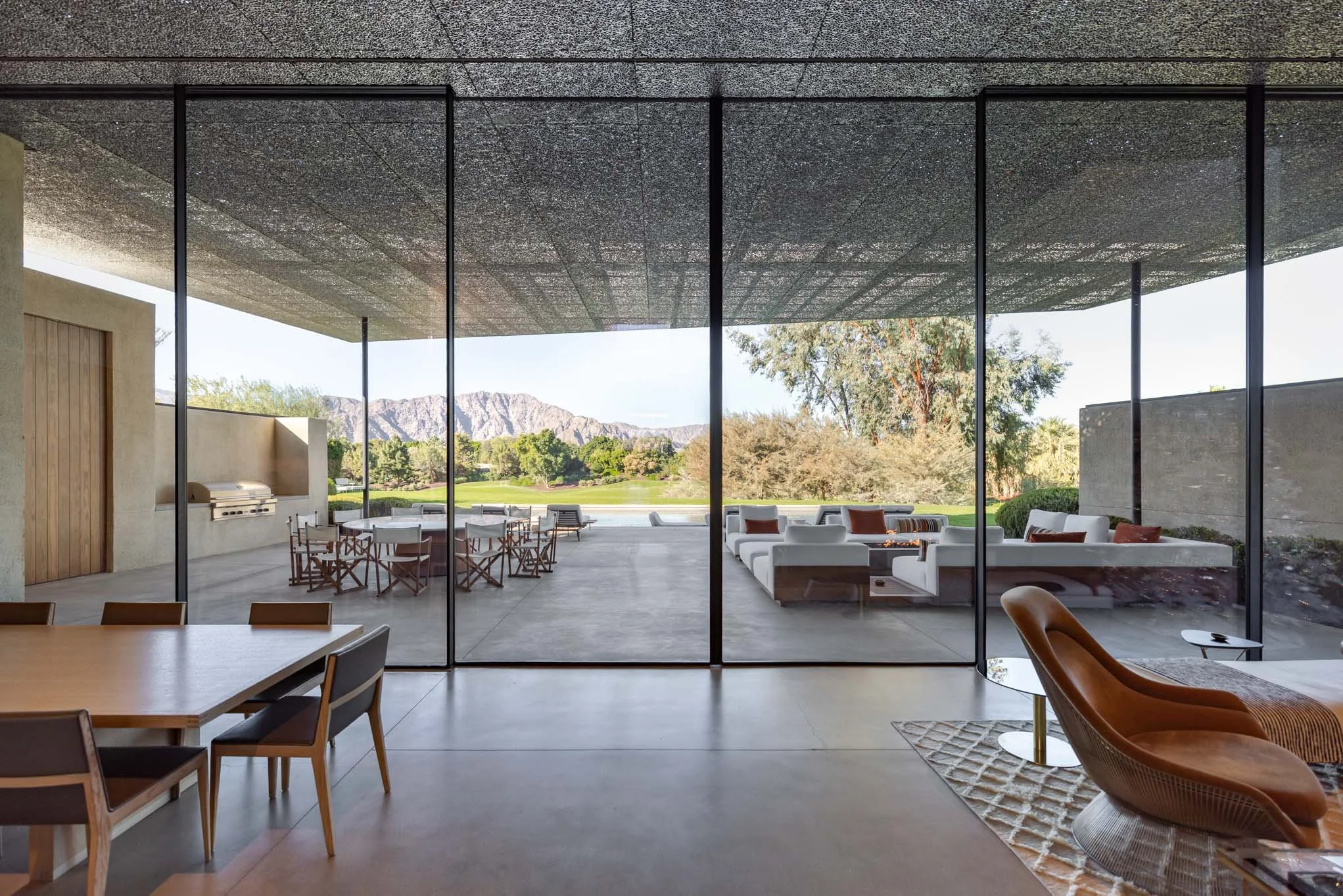 Indoor view of a modern living space with a large glass wall opening to an outdoor patio with seating, trees, mountains, and clear sky in the background.