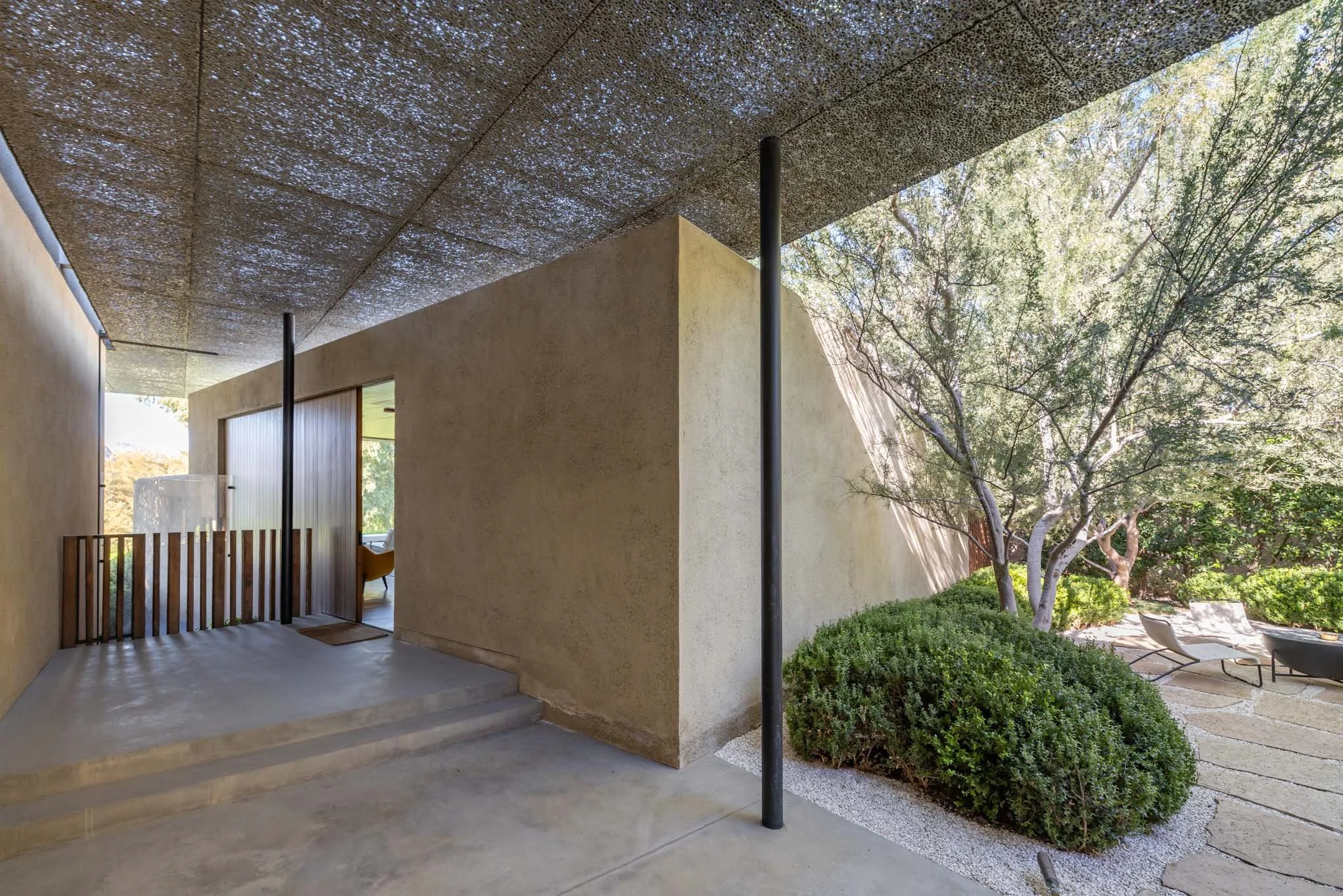 Modern house with a covered porch, showing textured ceiling, beige stucco walls, and an outdoor seating area surrounded by greenery and trees.