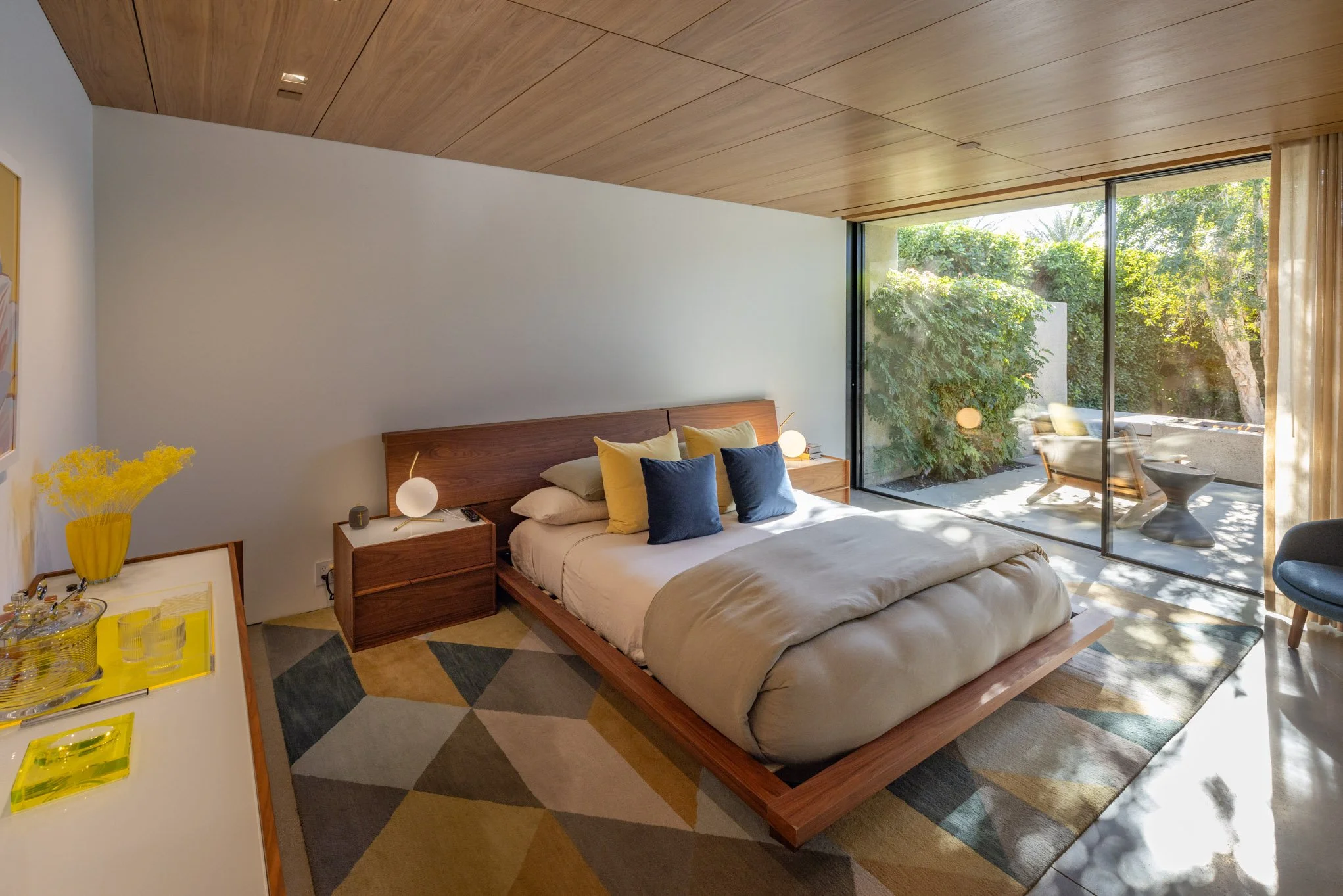 Modern bedroom with wooden ceiling and sliding glass door leading to outdoor seating area, queen bed with blue and yellow pillows, geometric rug, bedside lamps, and yellow decorative items.