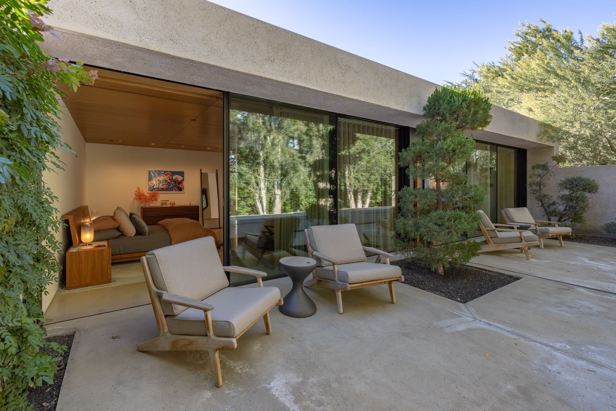 Modern house with large glass sliding doors, outdoor patio with chairs, small trees, and lush green trees in background.