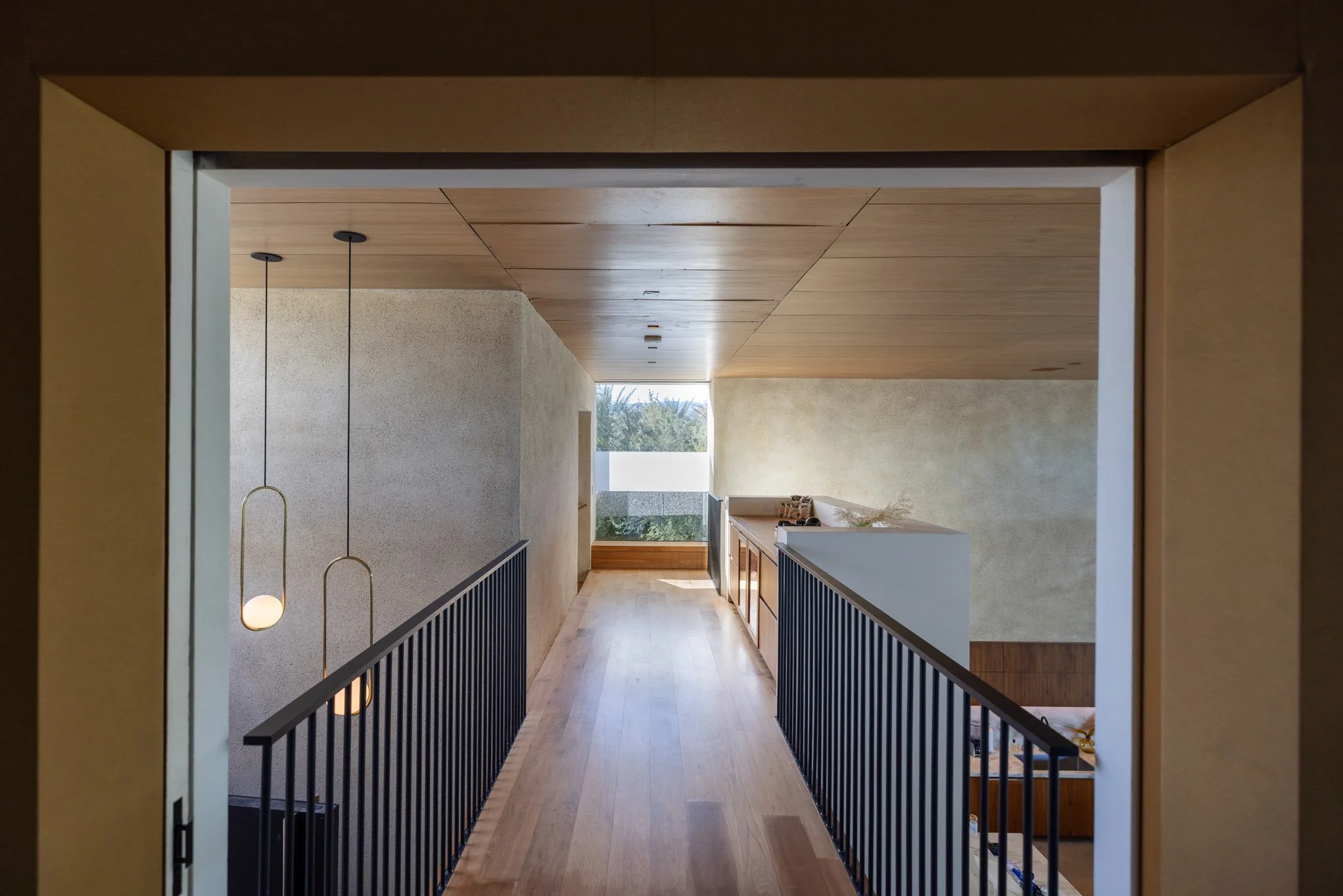 Interior view of a modern home corridor leading to a large window with trees outside, wooden flooring, black railing, and hanging pendant lights.