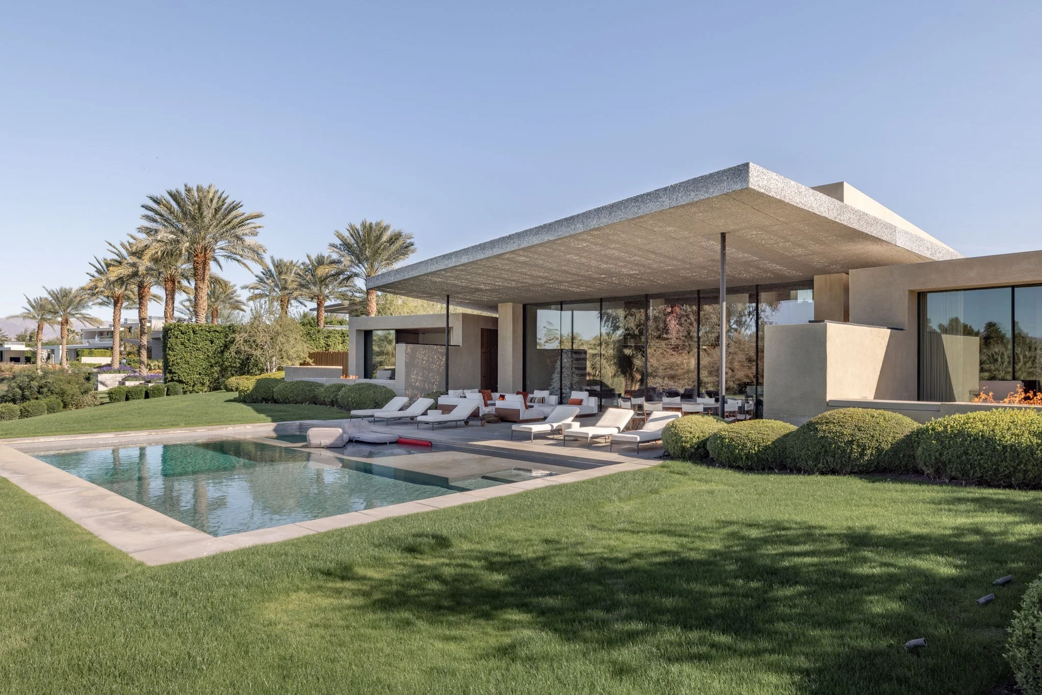 Modern house with large glass windows, outdoor lounge area with white chairs and couches, swimming pool with lounge chairs, palm trees, and green lawn under a clear sky.