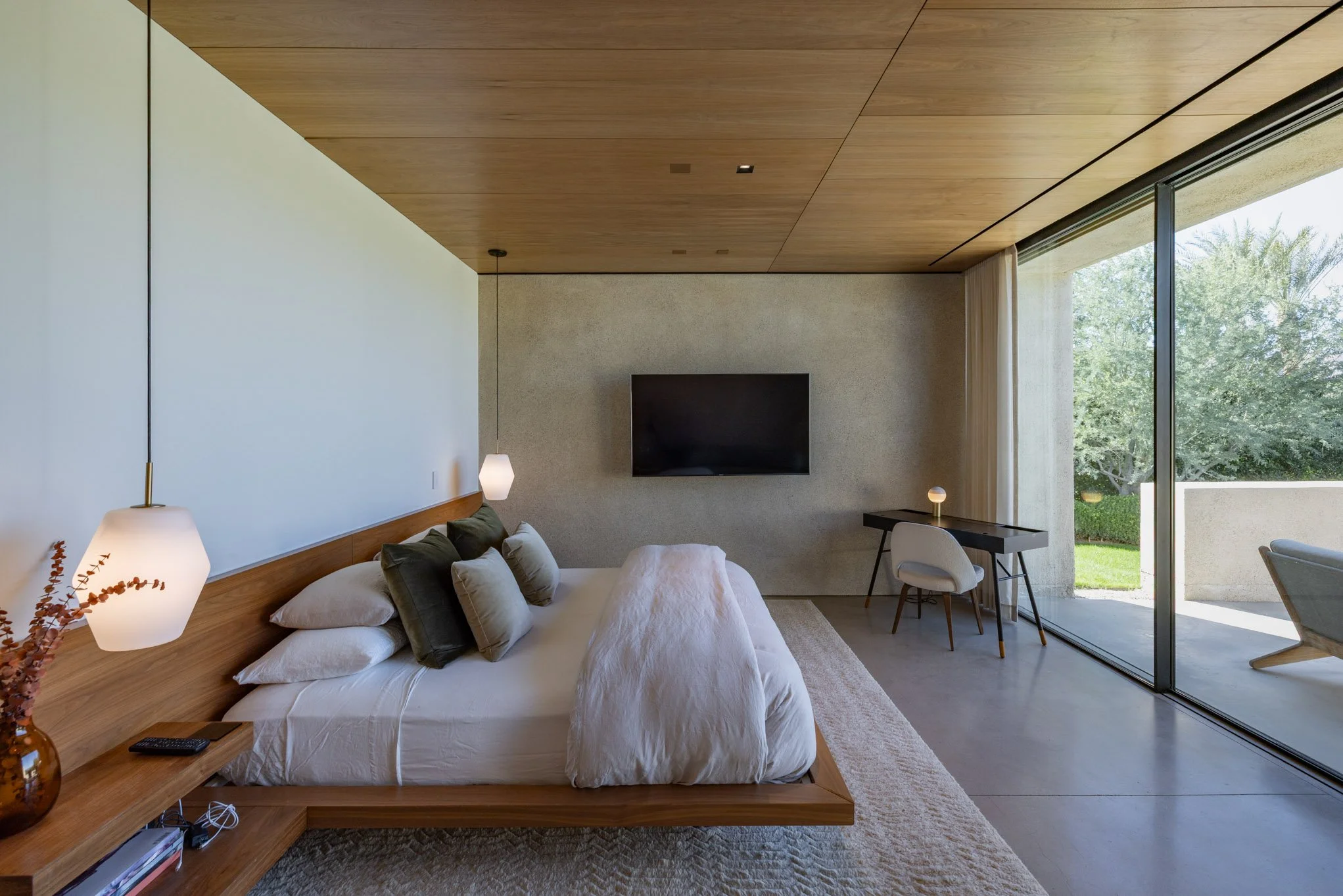 Modern bedroom with large glass sliding door to outdoor patio, minimalist decor, wooden ceiling, TV on textured wall, bed with multiple pillows, desk with chair, and hanging pendant lights.