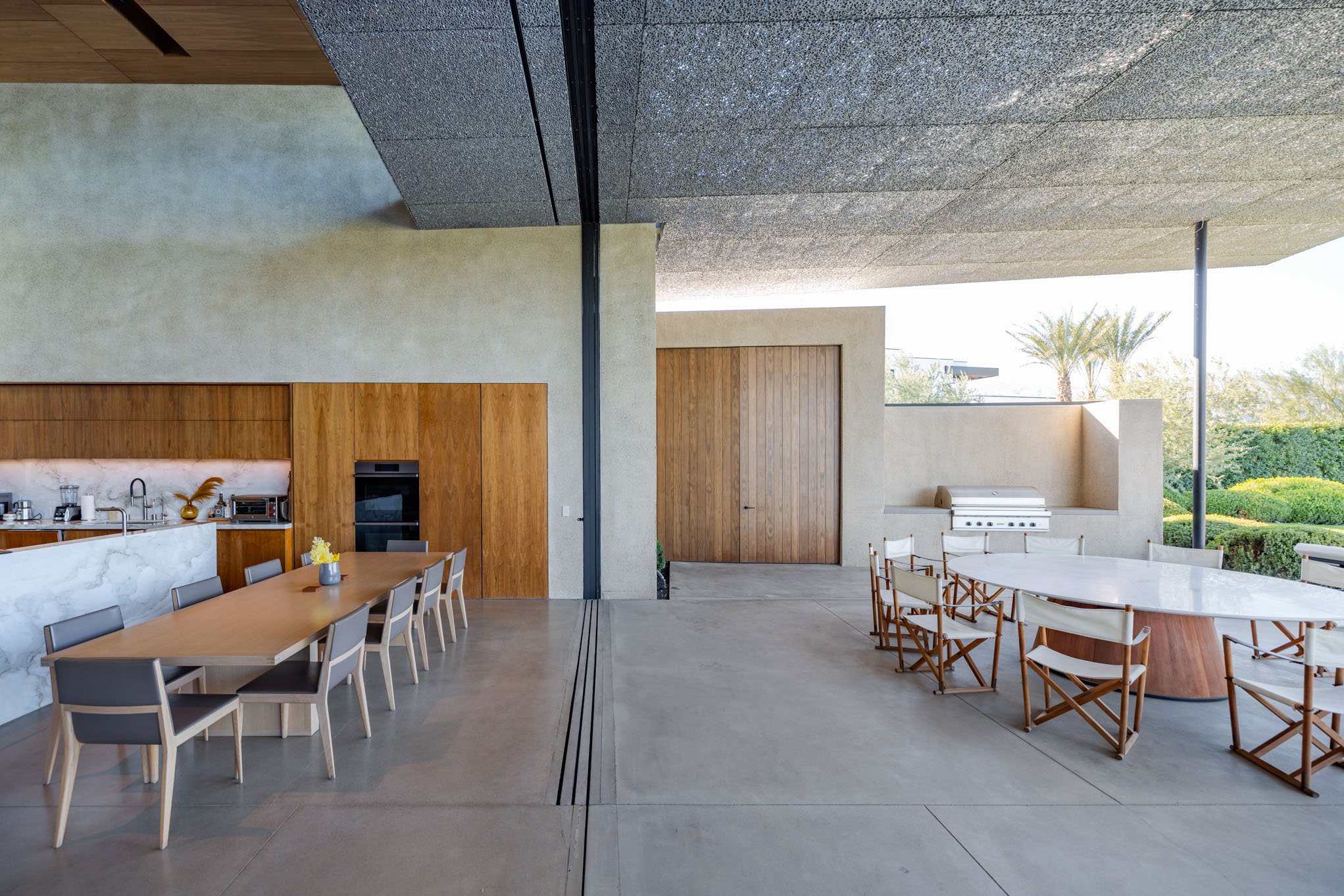 Modern indoor-outdoor kitchen and dining area with long wooden table, gray chairs, white marble countertop, built-in oven, outdoor grilling station, and surrounding greenery.