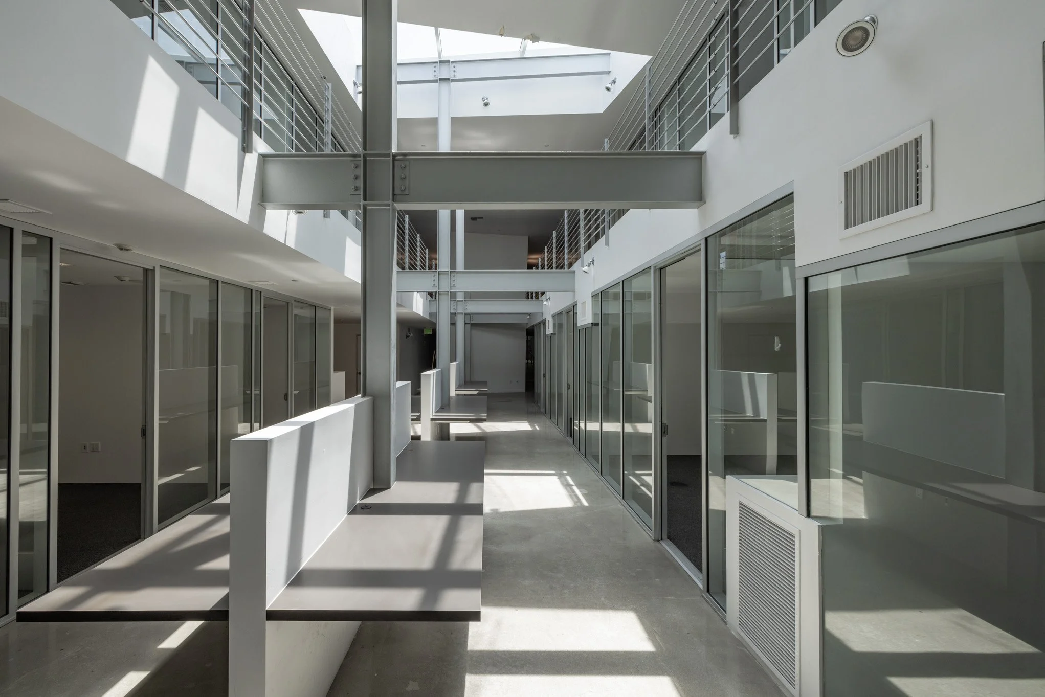 Modern interior corridor with glass walls, white walls, and metal beams, illuminated by natural light from skylights.