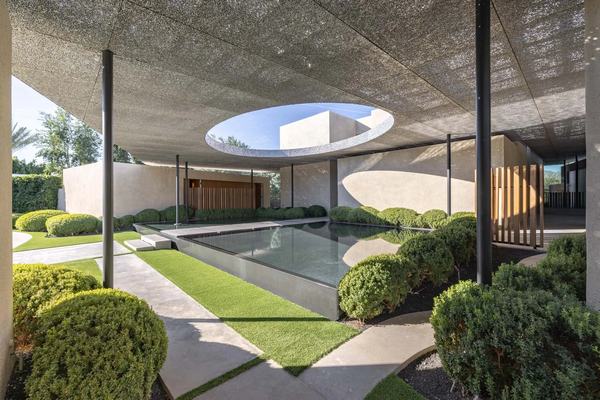 Modern architectural space with a reflective pool, surrounded by manicured bushes and greenery, under a large concrete ceiling with a circular opening revealing a blue sky.
