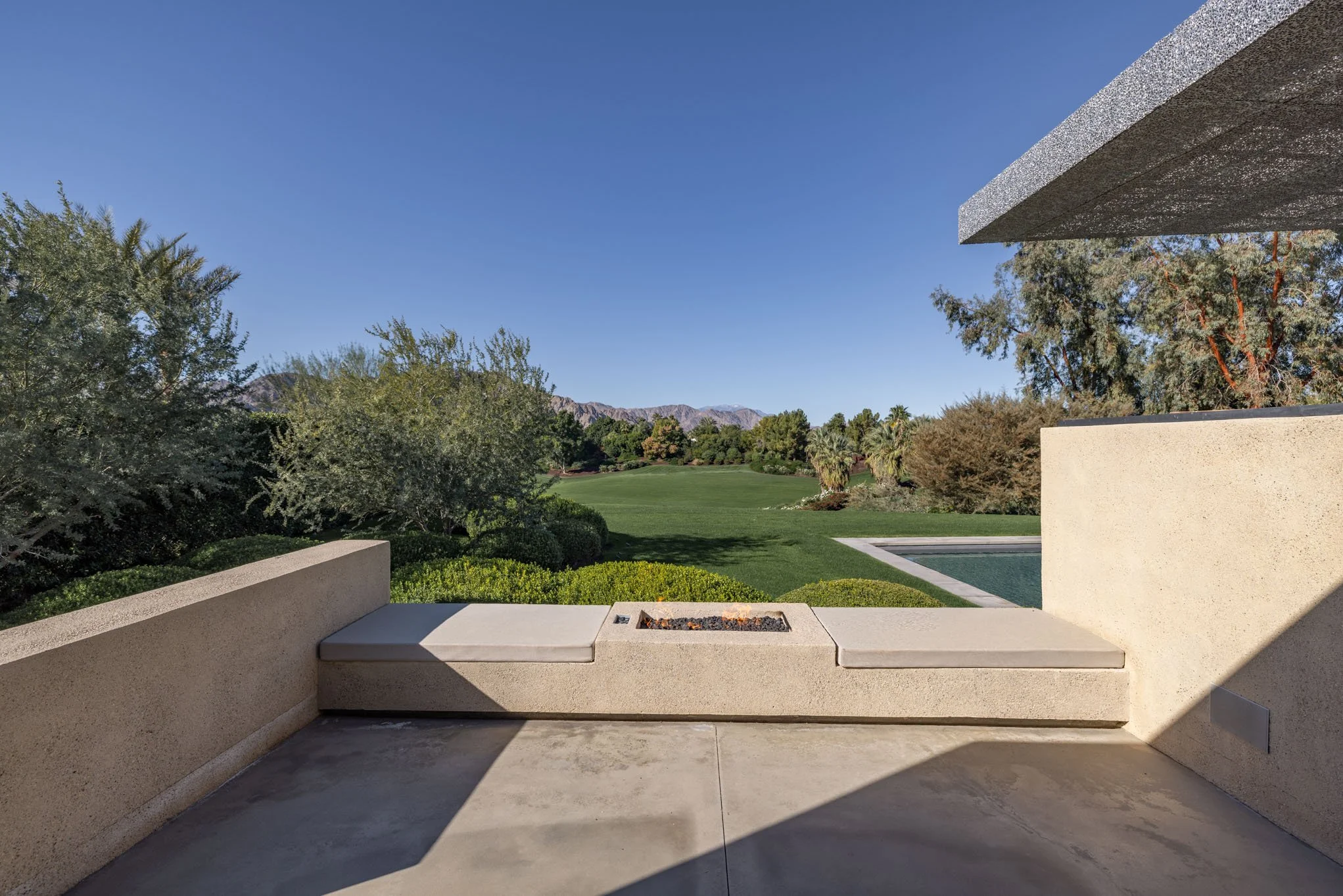 View from a modern outdoor patio with built-in seating and a fire pit, overlooking a green golf course with trees and mountains in the distance under a clear blue sky.