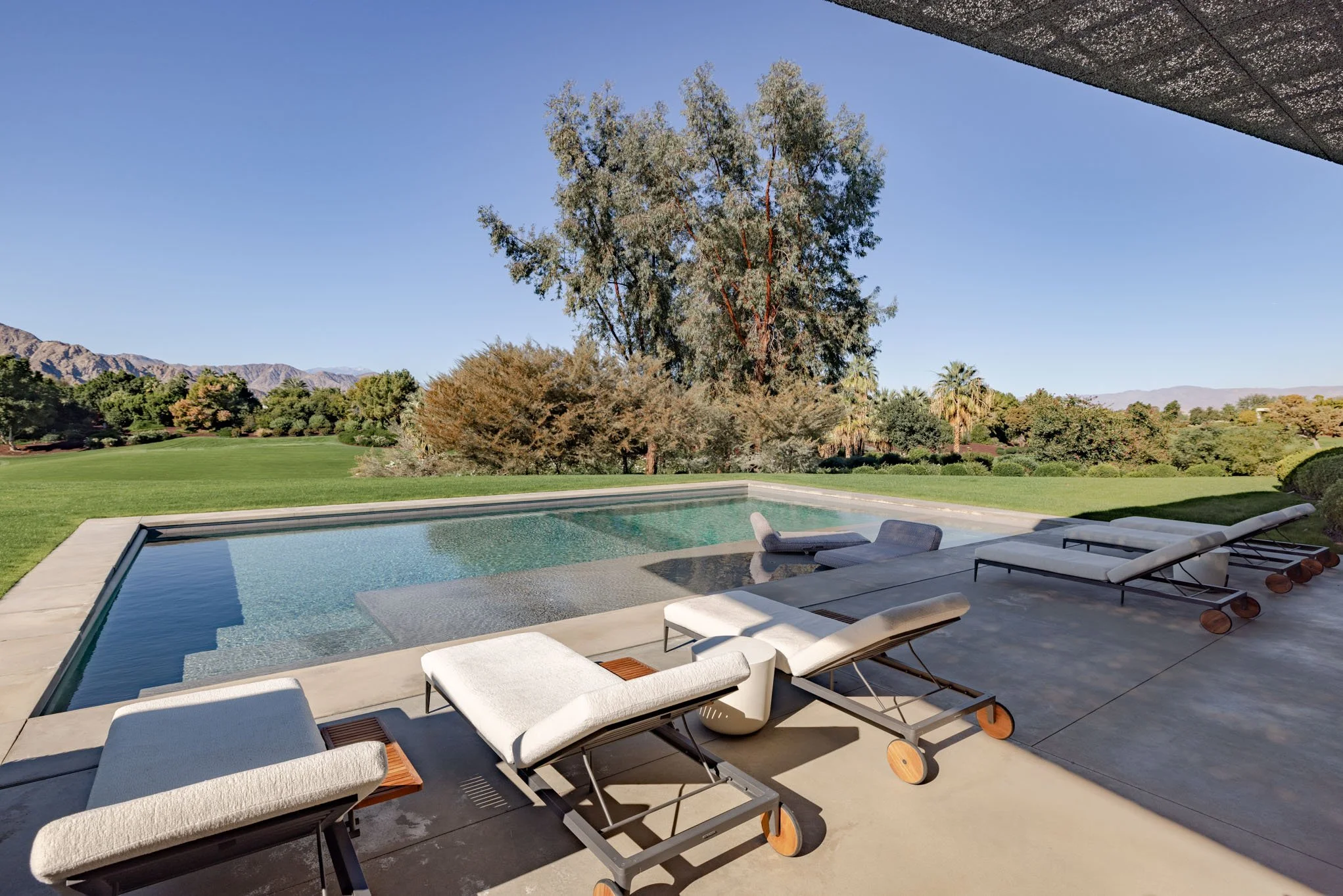 Luxury backyard with a modern swimming pool, four white lounge chairs, and a lush tree-filled landscape with mountains in the background under a clear blue sky.
