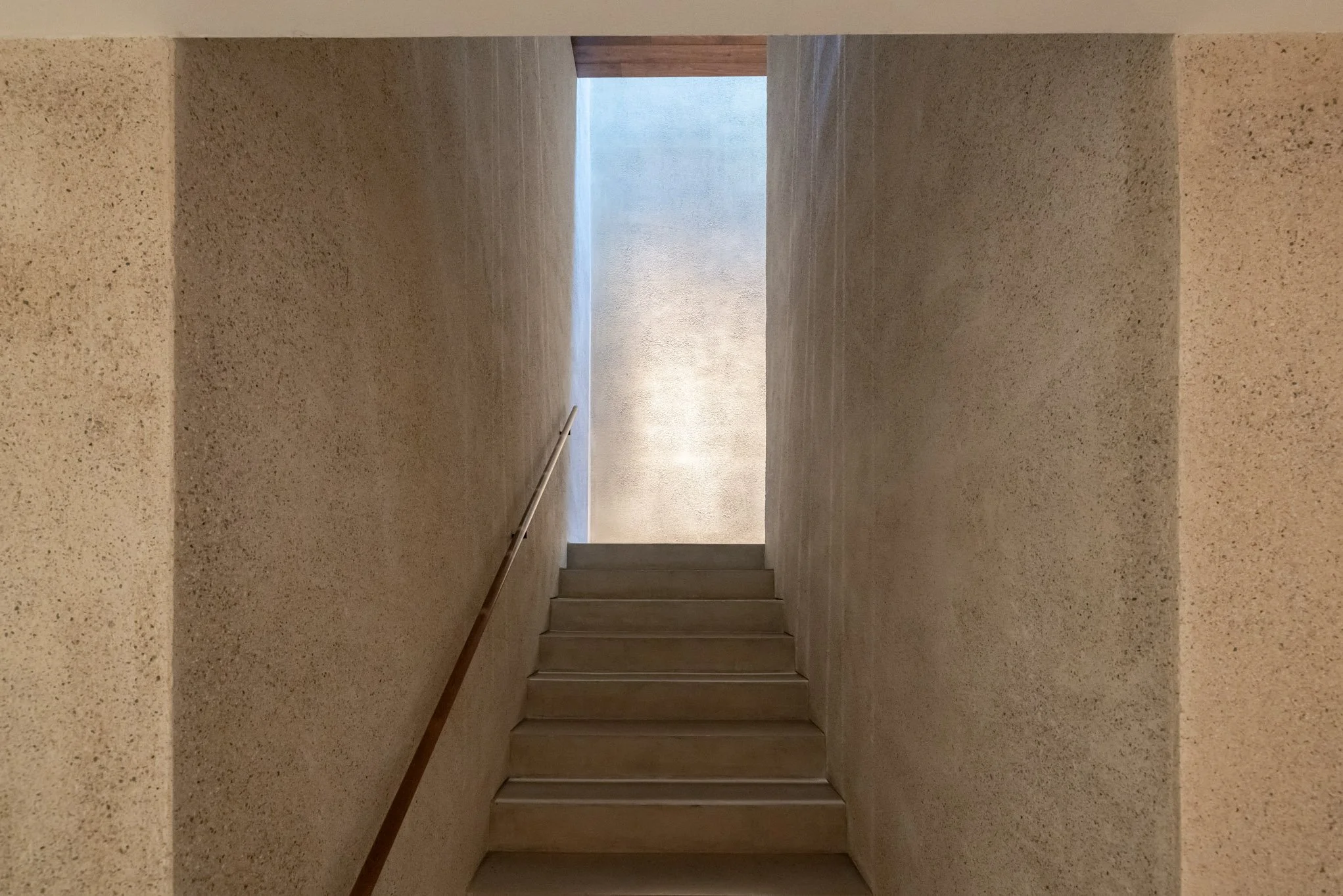 A staircase with beige walls and a metal handrail leading up to a narrow opening with natural light and blue sky visible above.