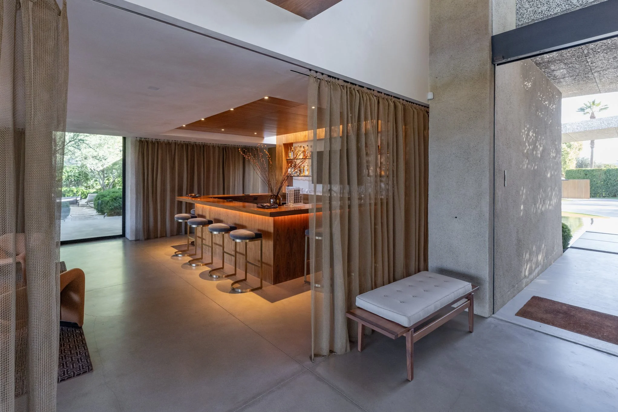 Modern indoor lounge with a wooden bar counter, high stools, and large glass sliding doors leading to an outdoor patio with greenery.