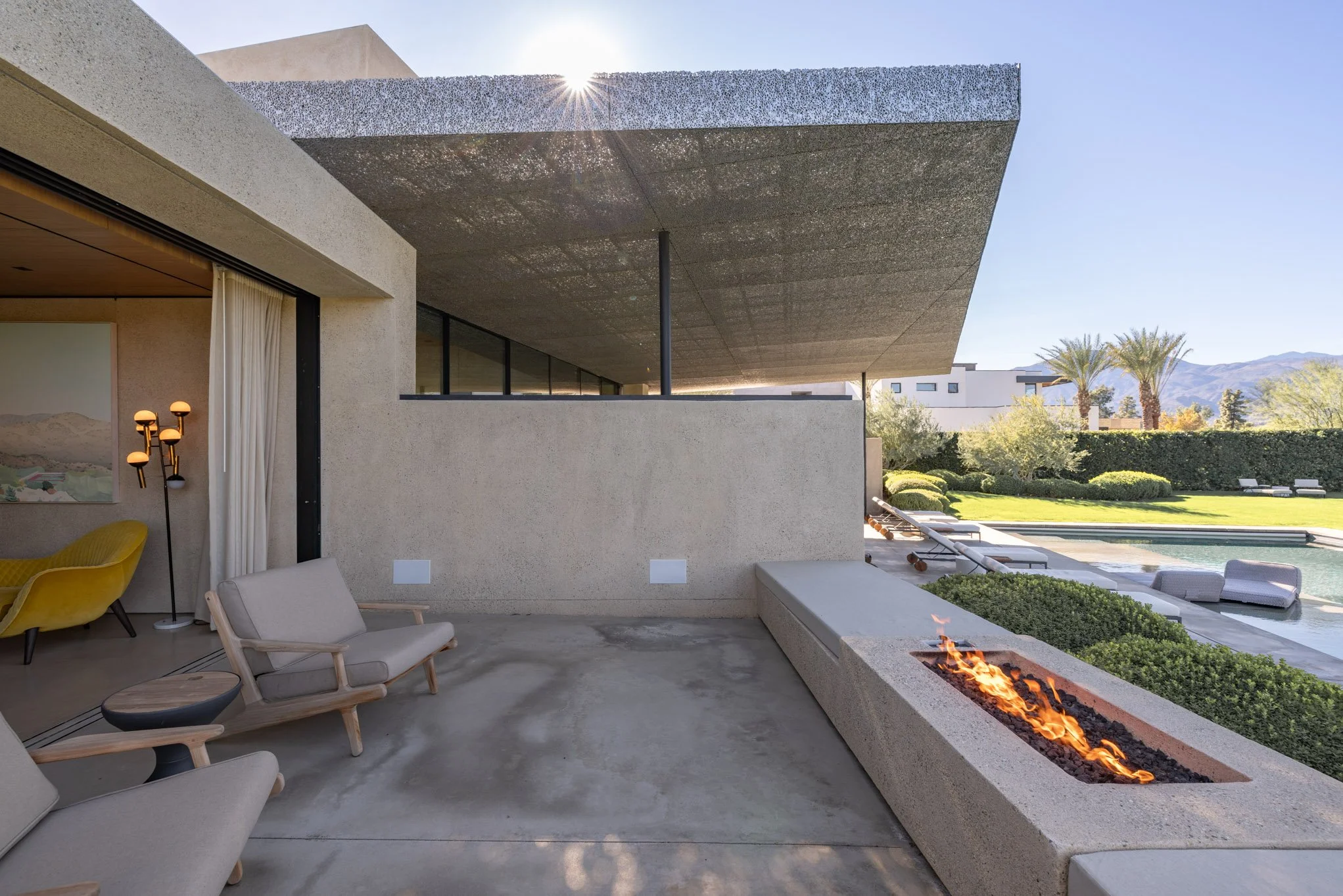 Modern outdoor patio with fire pit, lounge chairs, and scenic yard with pool, palm trees, and mountains in the background.
