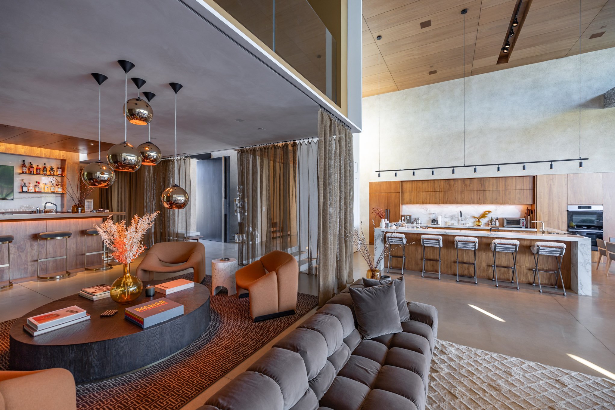 Modern open-concept living space featuring a kitchen with a marble island and bar stools, a seating area with a brown leather sofa and armchairs, a round coffee table with books and decorative items, a bar area with bottles and glasses, and large win