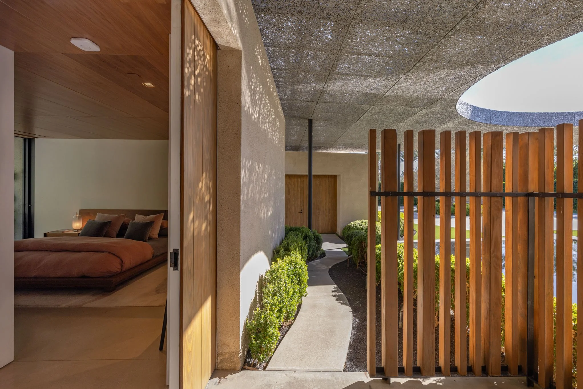 View from inside a modern house showing a wooden door opening to a concrete pathway bordered by green bushes, with a wooden fence and garden outside, and a bedroom with a bed and pillows visible through the open door.