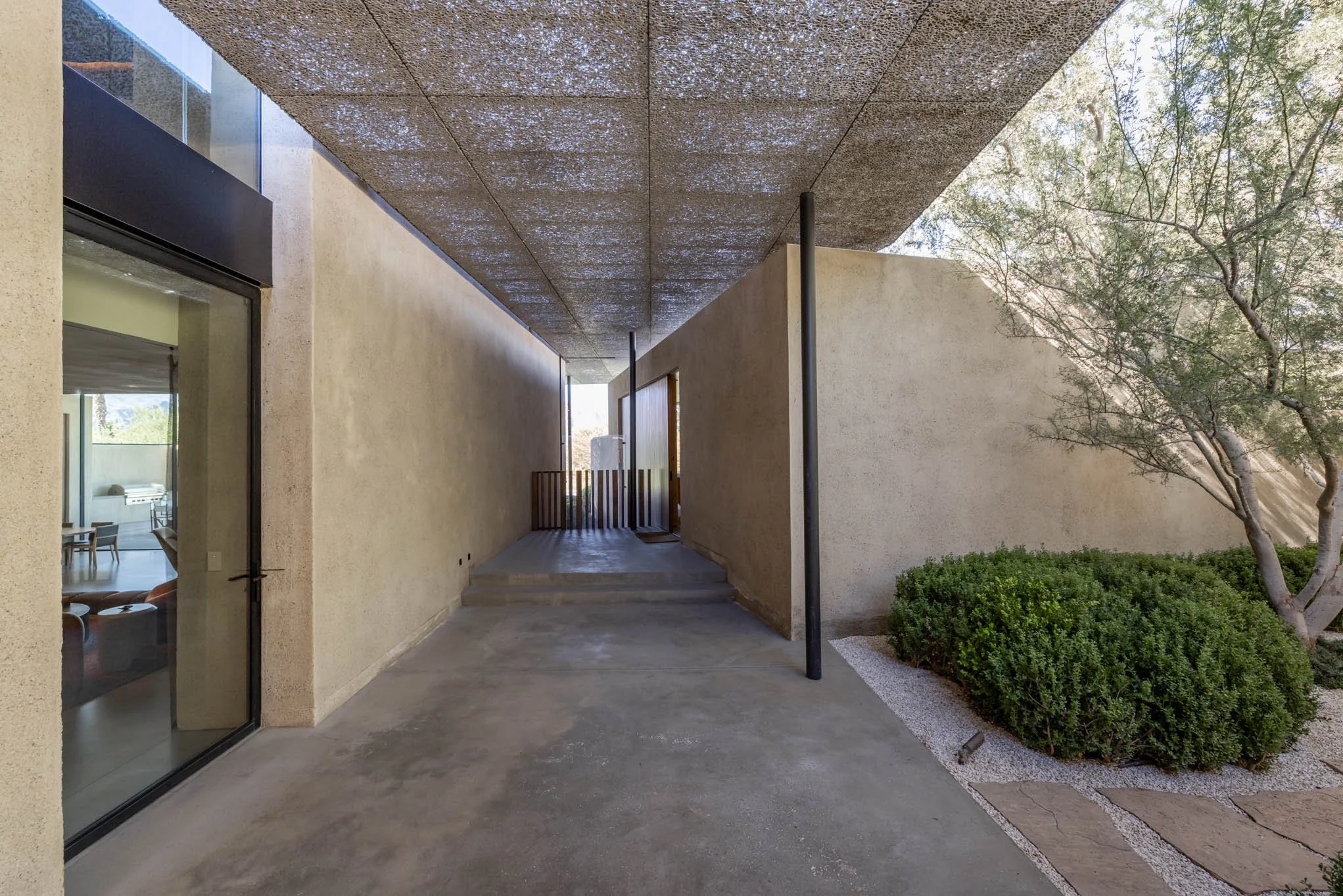 Outdoor corridor of a modern building with concrete flooring, beige stucco walls, and a textured ceiling. There are steps leading up to a wooden gate at the end of the corridor. To the left, there is a large glass door revealing a living room with fu
