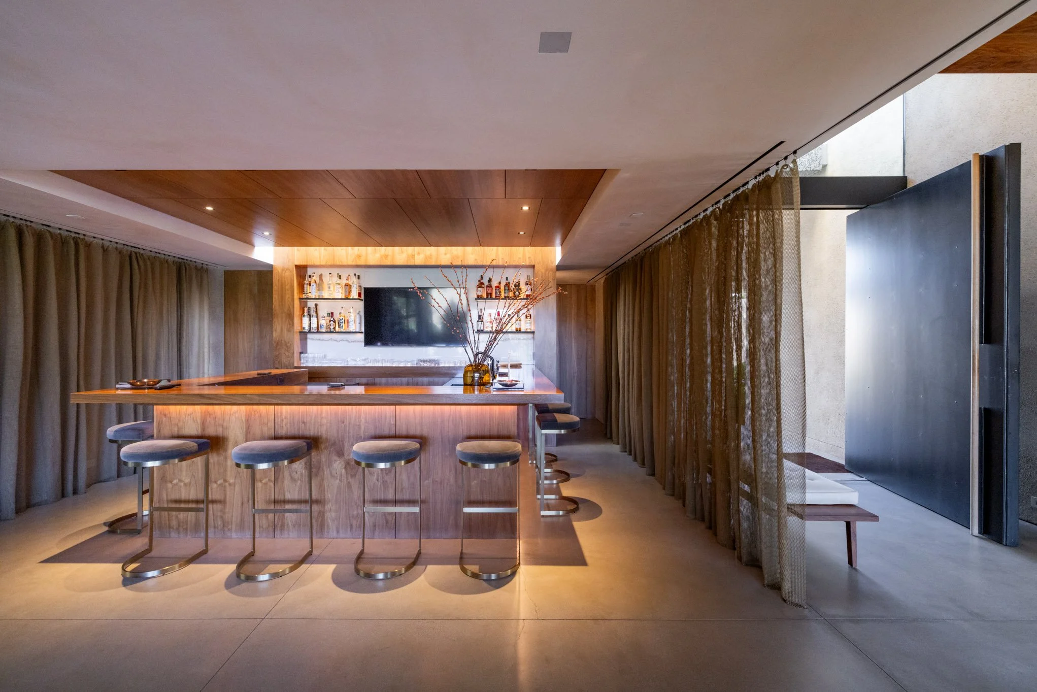 Modern indoor bar with wooden counter, stools, shelves of liquor bottles, a TV, and beige curtains dividing the space.