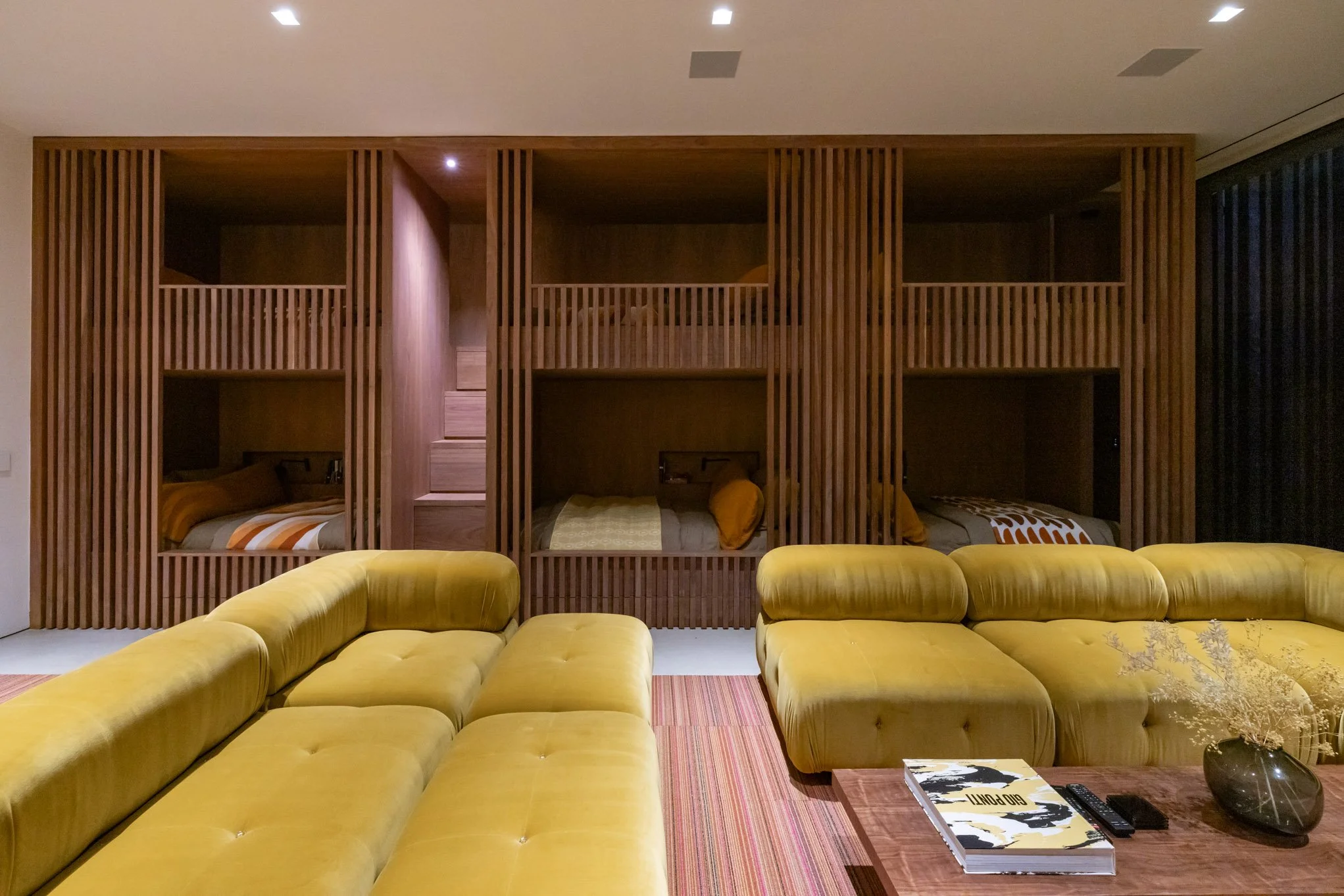 Modern living room with a yellow sectional sofa, a wooden coffee table, and built-in wooden bunk beds in the background.