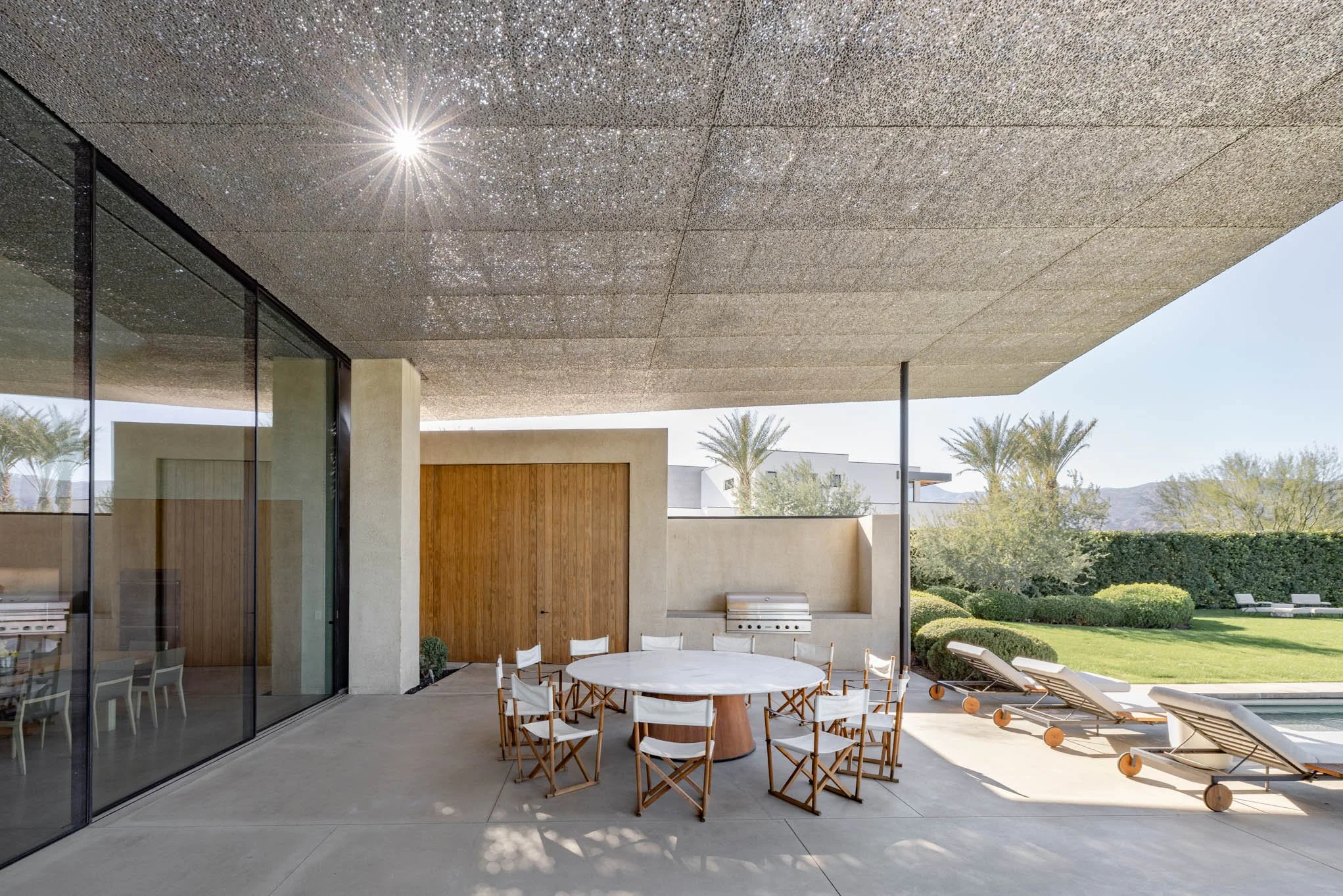 Modern outdoor patio with round dining table and chairs, lounge chairs by the pool, surrounded by lush greenery and palm trees, under a large overhanging ceiling.