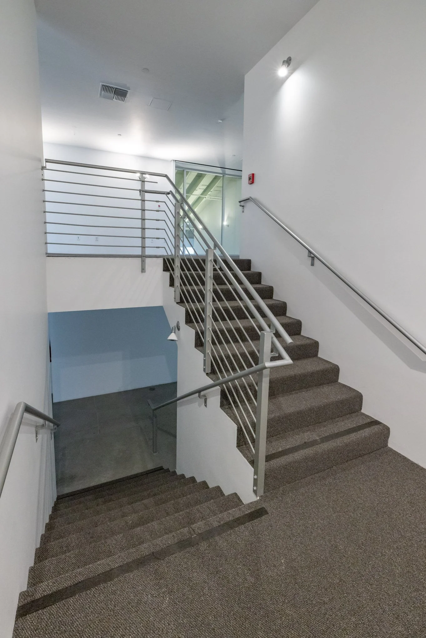 Interior view of a staircase with metal handrails leading down to a lower level in a modern building with white walls and carpeting.