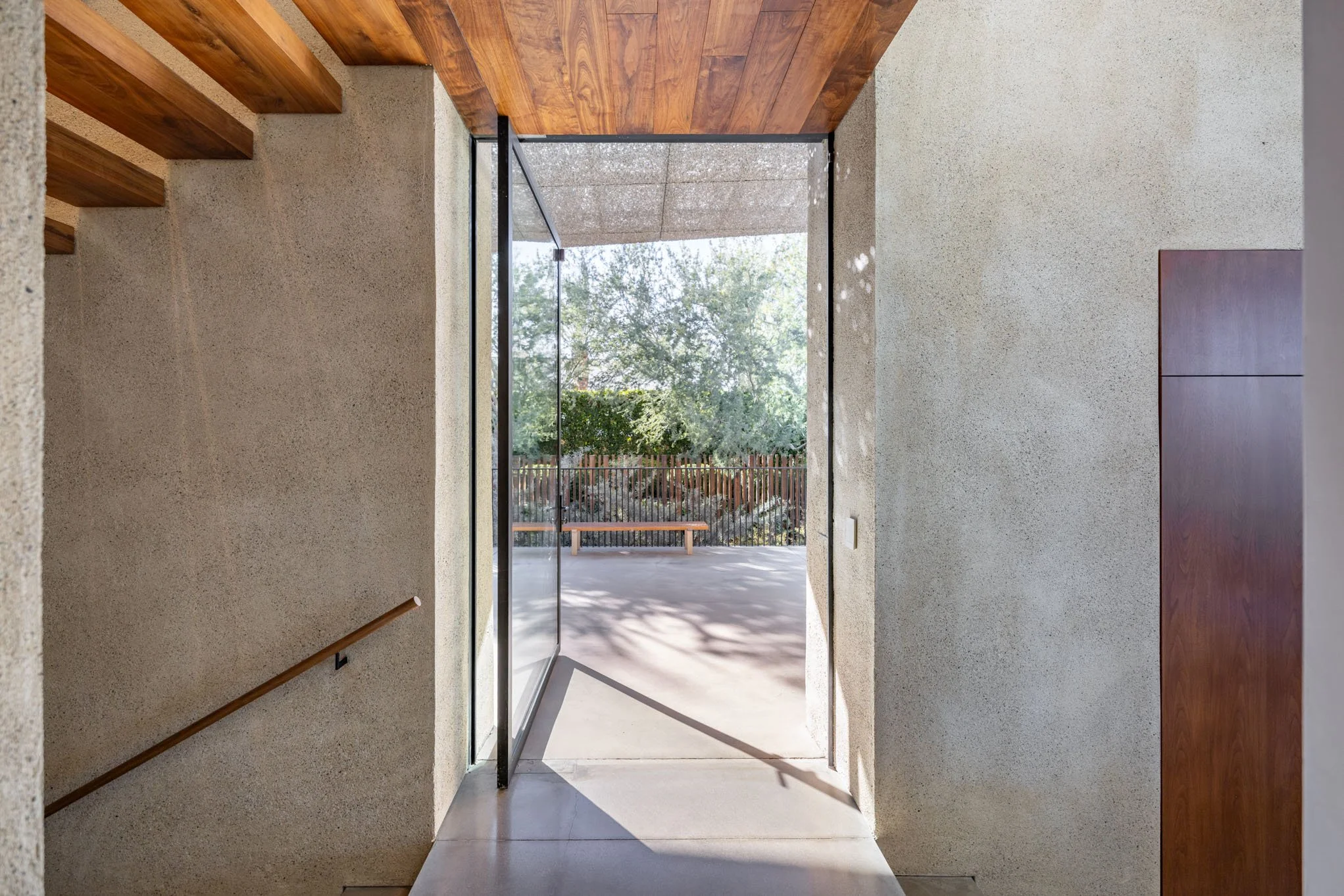 Interior view of a modern building entrance with a concrete wall, wooden ceiling, and an open glass door leading outside to a patio area with a bench and trees.