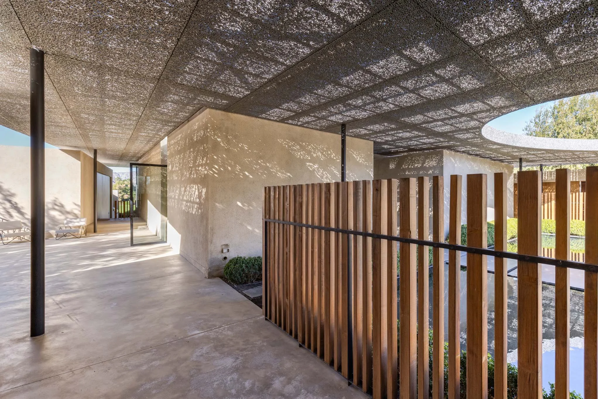 Modern outdoor space with concrete flooring, a textured black ceiling, a beige wall with shadows, a wooden fence, and a small pond visible through the fence.
