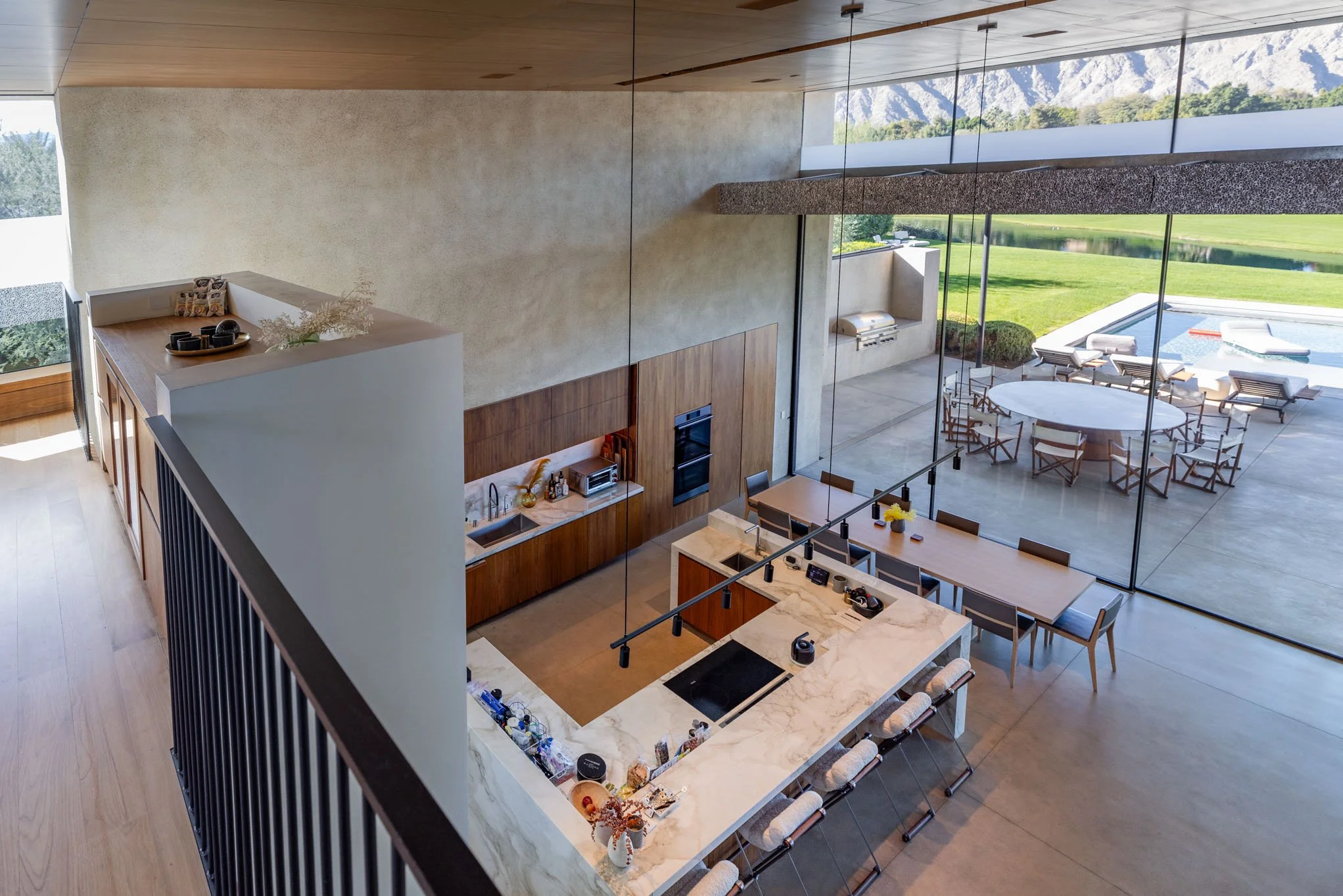 Modern open-concept kitchen with marble island, wooden cabinetry, large glass windows revealing an outdoor patio with a pool and lounge area, mountain and green landscape in the background.