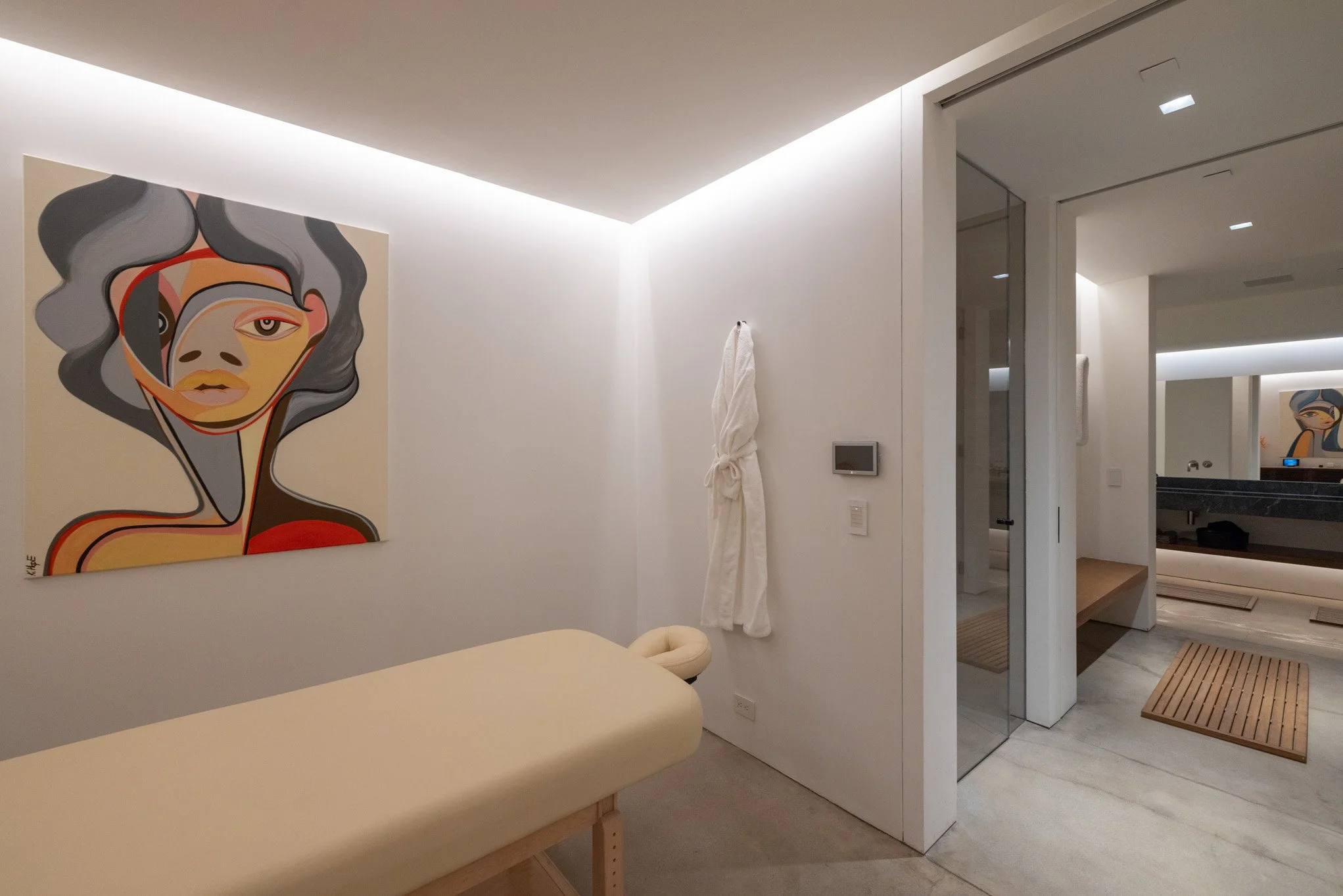 Modern interior room with white walls and ceiling, a colorful abstract portrait painting of a woman's face on the left wall, a massage or examination table in the foreground, a white robe hanging on the wall, a digital control panel, and a sliding gl
