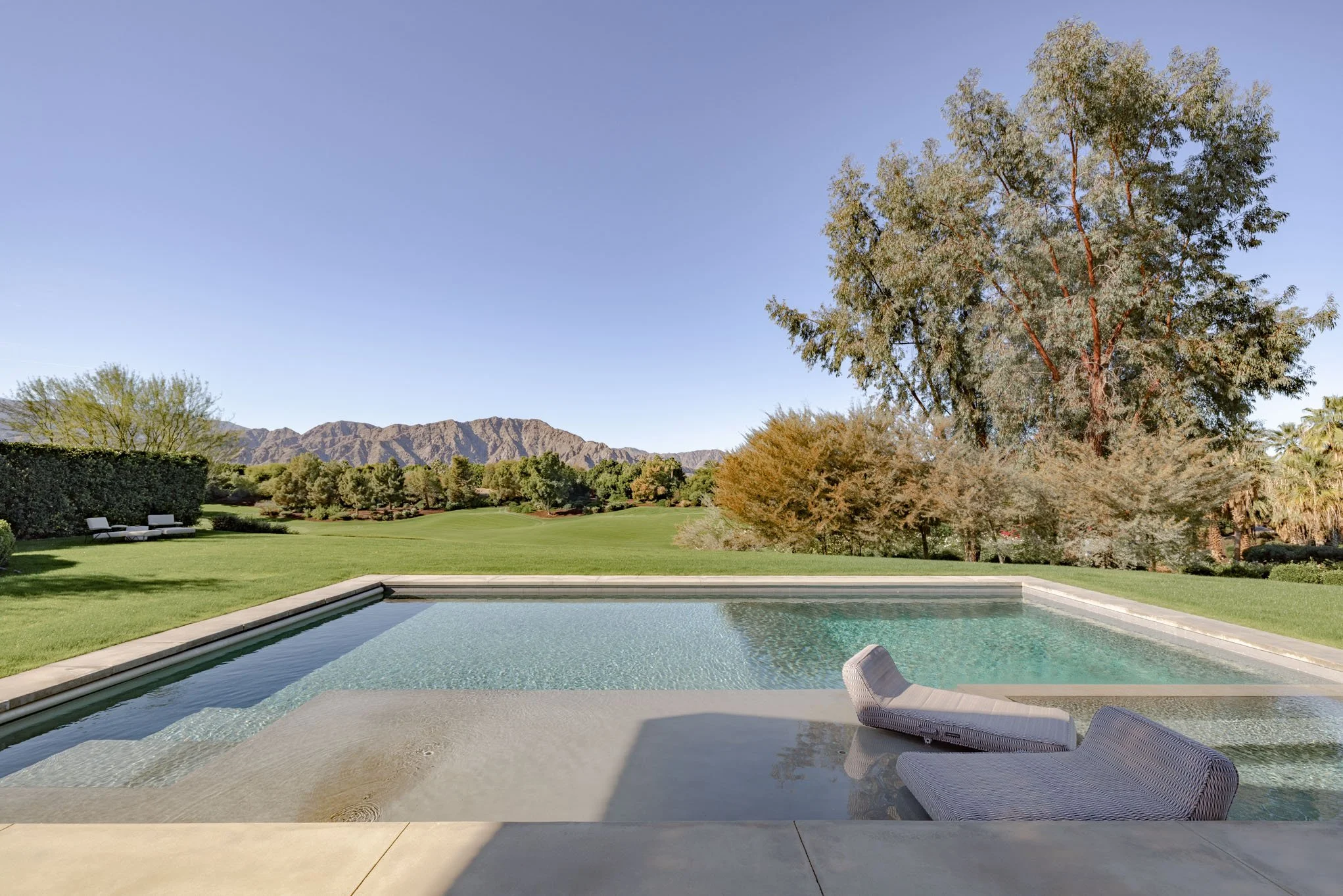 A backyard with a rectangular swimming pool, lounge chairs, trees, green lawn, mountain range in the background, and clear blue sky.
