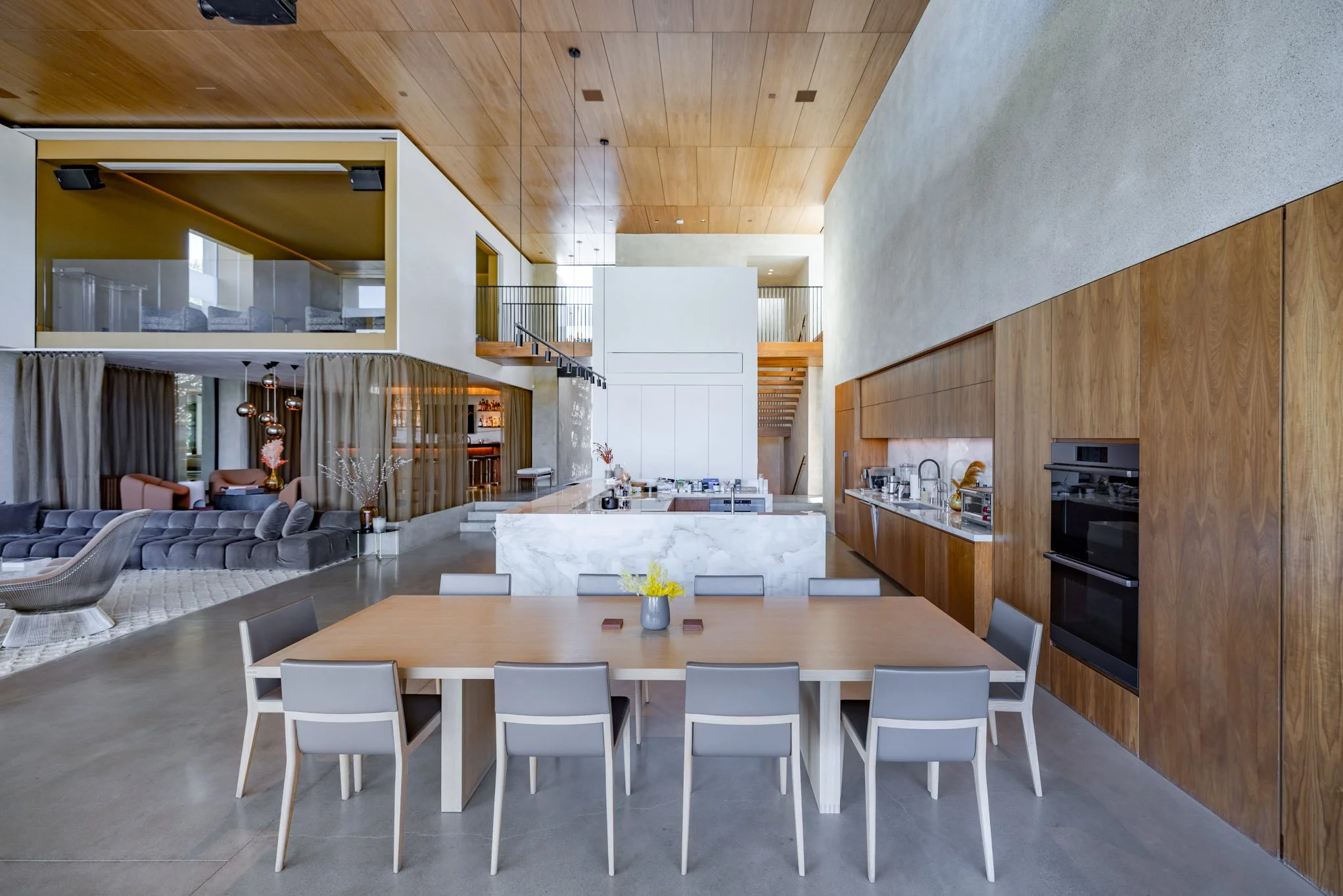 Modern open-concept kitchen and dining area with wooden accents, marble island, and a view into a living room with a sofa and hanging pendant lights.