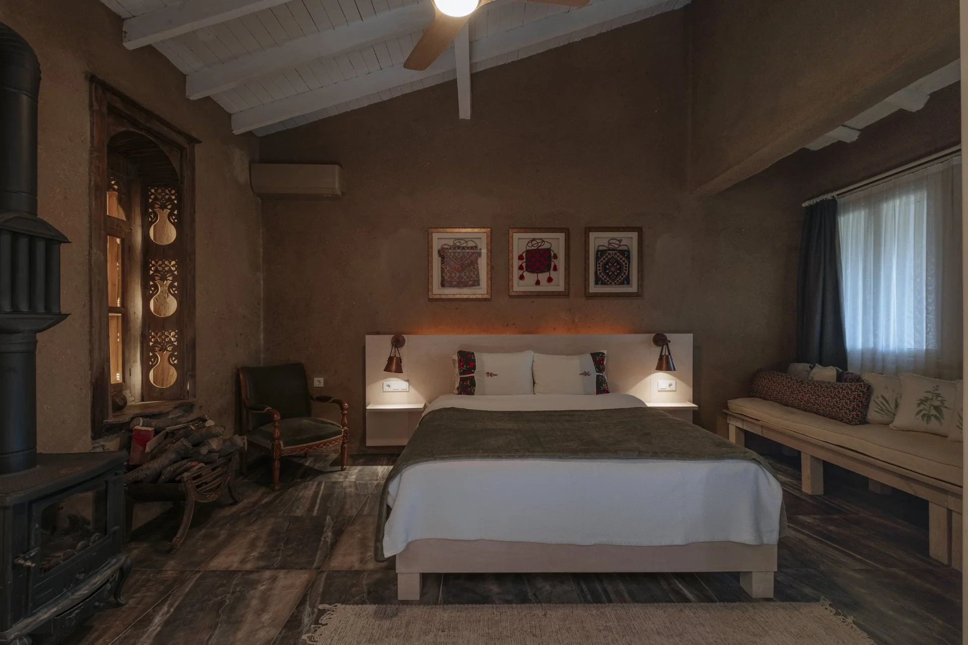 Rustik tarzda yatak odası, ahşap detaylar, karışık renkli yorgan ve yastıklar, duvarda üç parça sanat eseri, yanlarda lamba ışıkları, pencere ve perdeler, klima ve şömine.