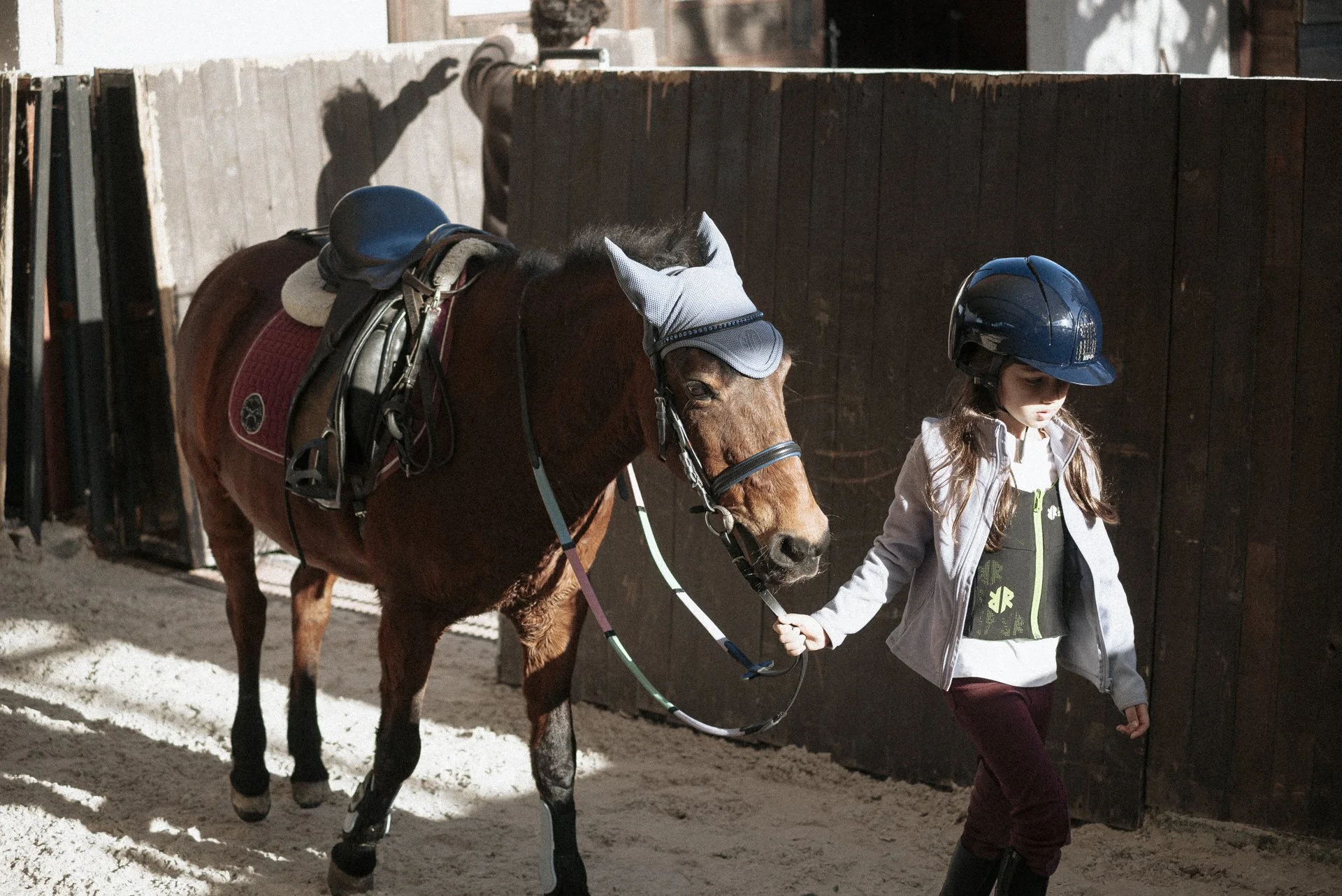 Genç bir kız binicilik kıyafetleriyle, bir atı tutarak binicilik eğitimi yapıyor, atın üzerinde yelek ve kask var.