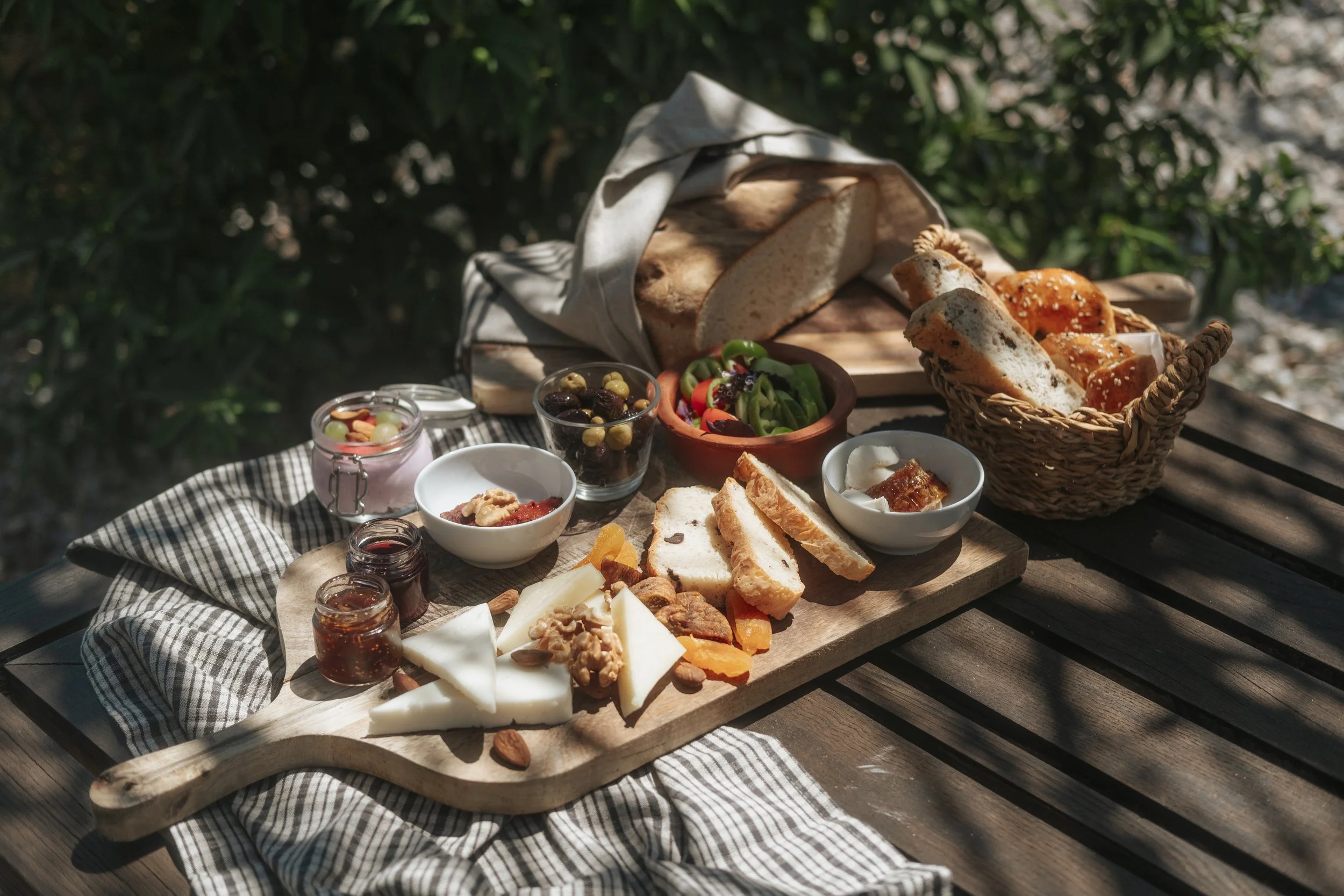 Kahvaltı tabağı, peynir, zeytin, ekmek, reçel, meyve ve çeşitli yiyecekler içeriyor. Masa açık havada, doğada keyifli bir ortamda.