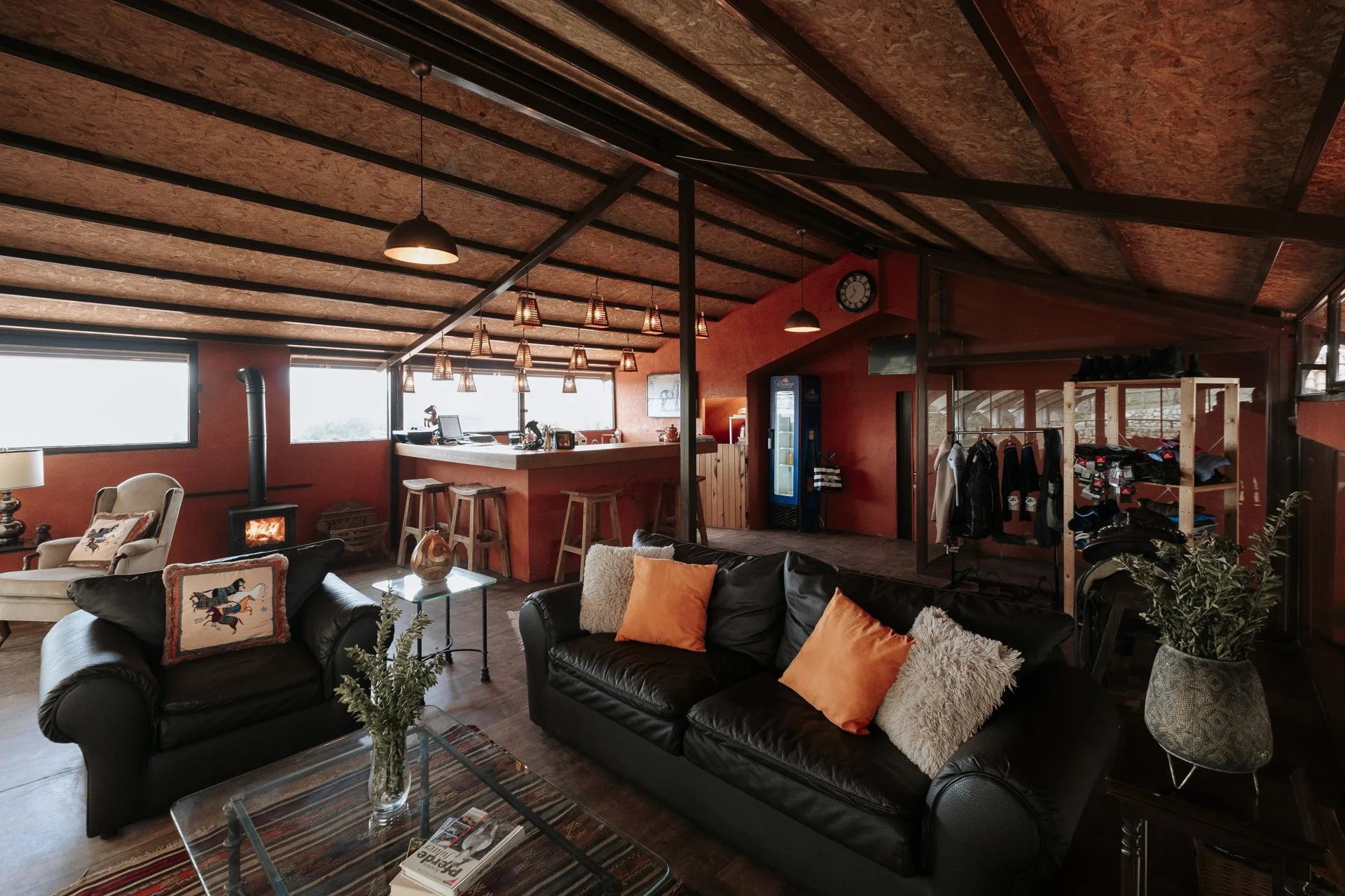 Kırmızı duvarlara ve ahşap tavanlı oturma odası. Çeşitli koltuklar, yastıklar ve bir sehpa var. Pencere ve bar alanı görüntüleniyor.