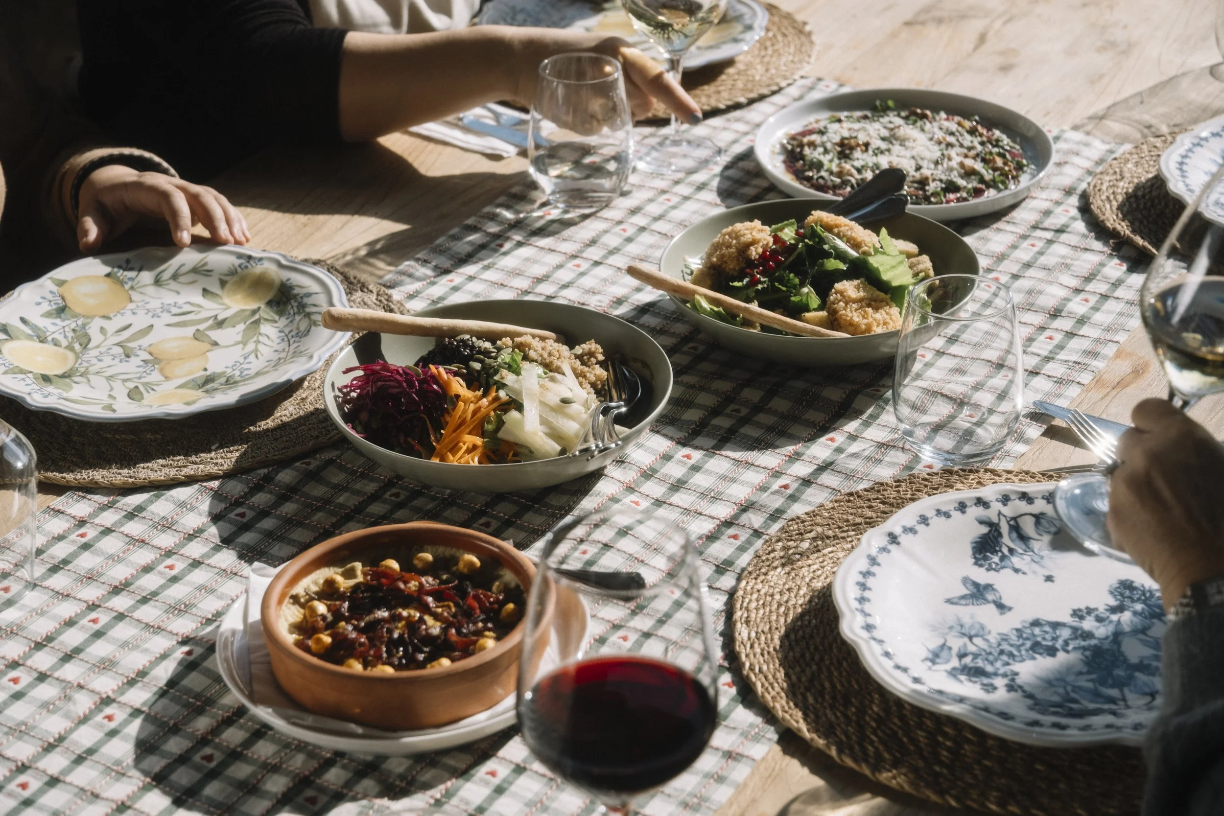 Bir masa üzerinde çeşitli tabaklarda salata, humus ve şarap bulunuyor, masa örtüsü kısıtlı ve masa etrafına oturmuş kişiler var.