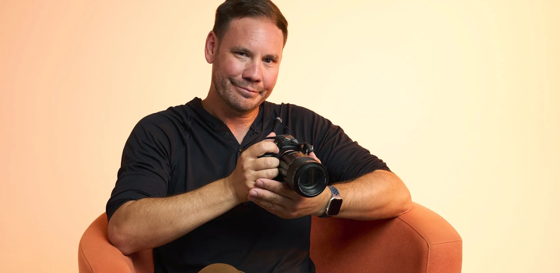 Jason Collin, Pacific Grove, CA personal branding photographer, holding a professional camera while seated in an orange chair in his photography studio.