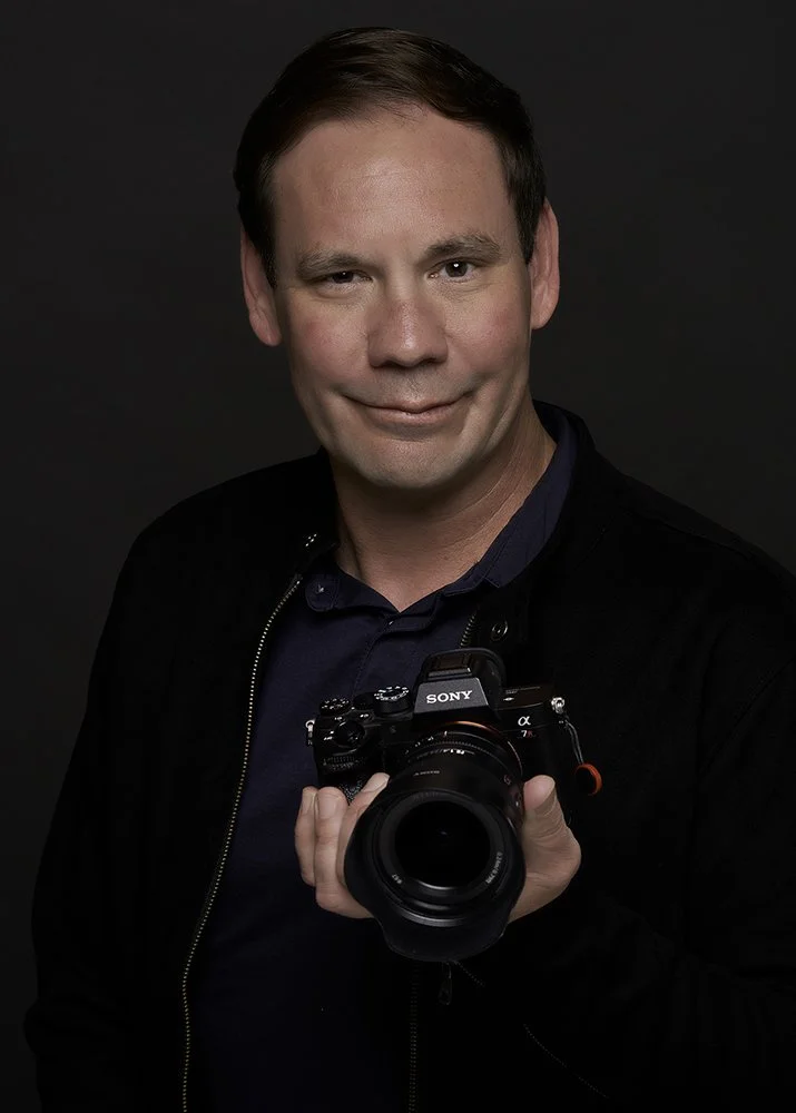 Photography Jason Collin holding a camera photographed on a gray background in his Pacific Grove, CA studio.