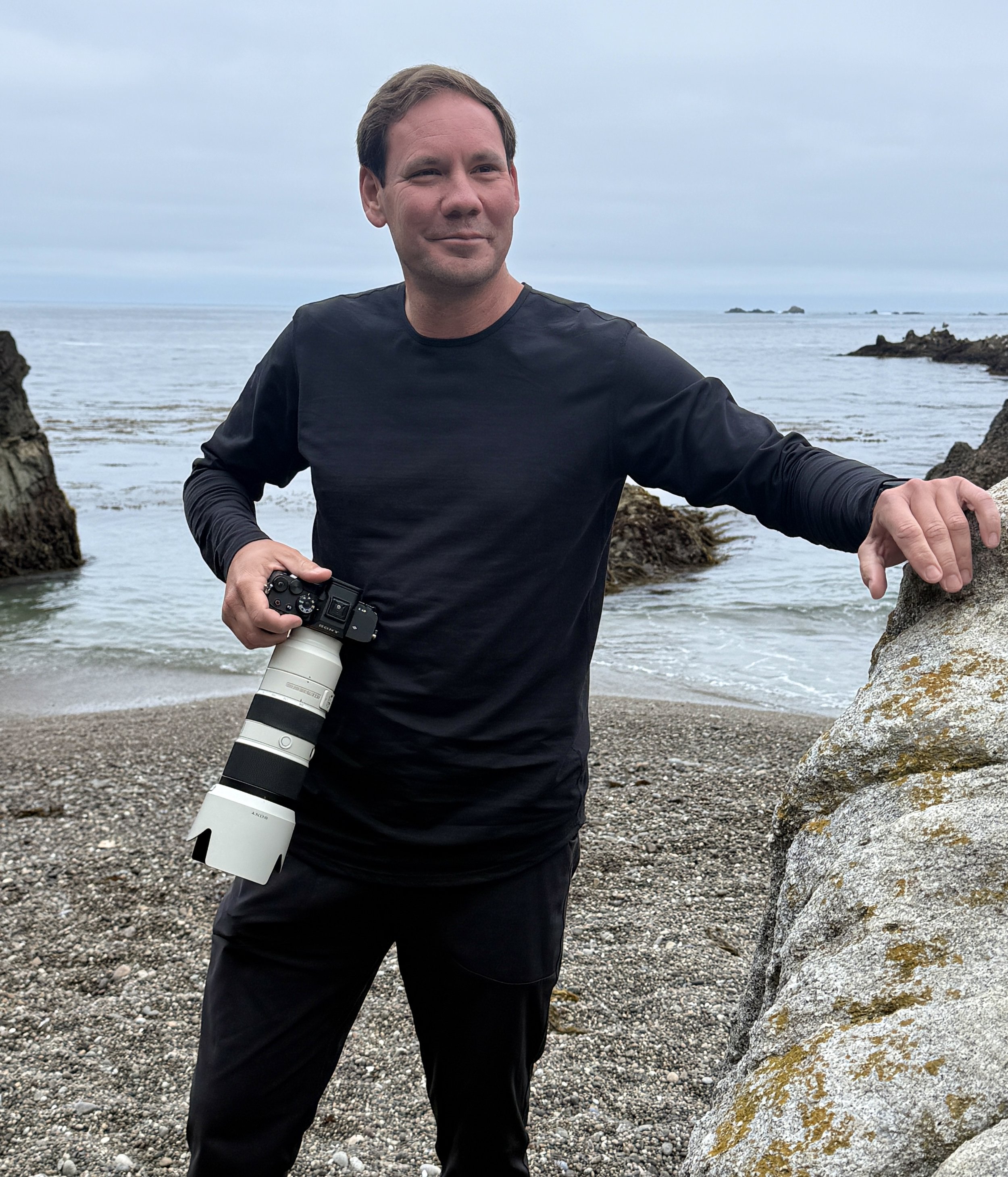 Photographer Jason Collin standing along the Pacific Grove coast with camera in hand, Monterey Bay in the background