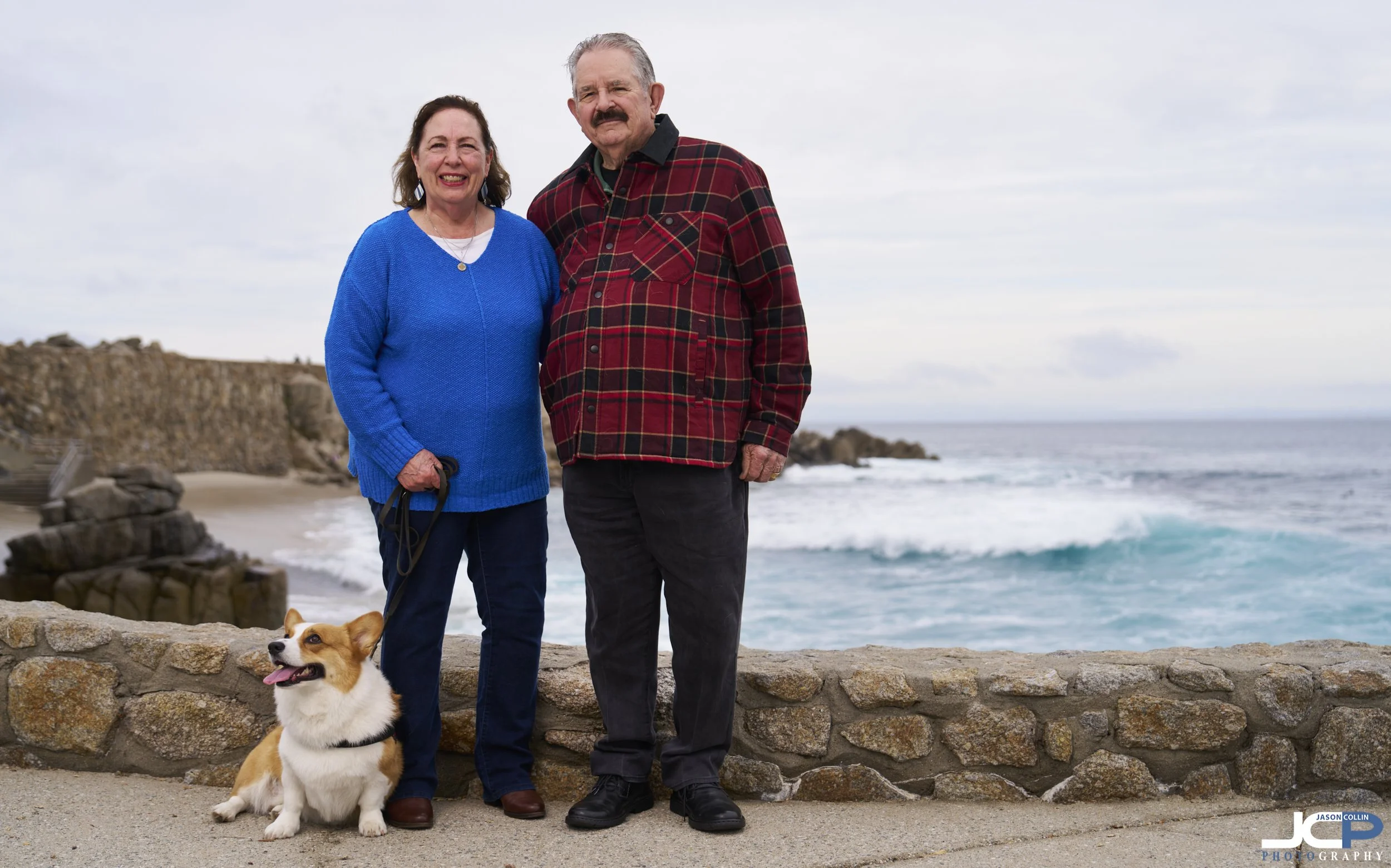 Couple with their corgi dog standing together at Lover’s Point Park in Pacific Grove, California with ocean waves in the background during a relaxed family portrait session.