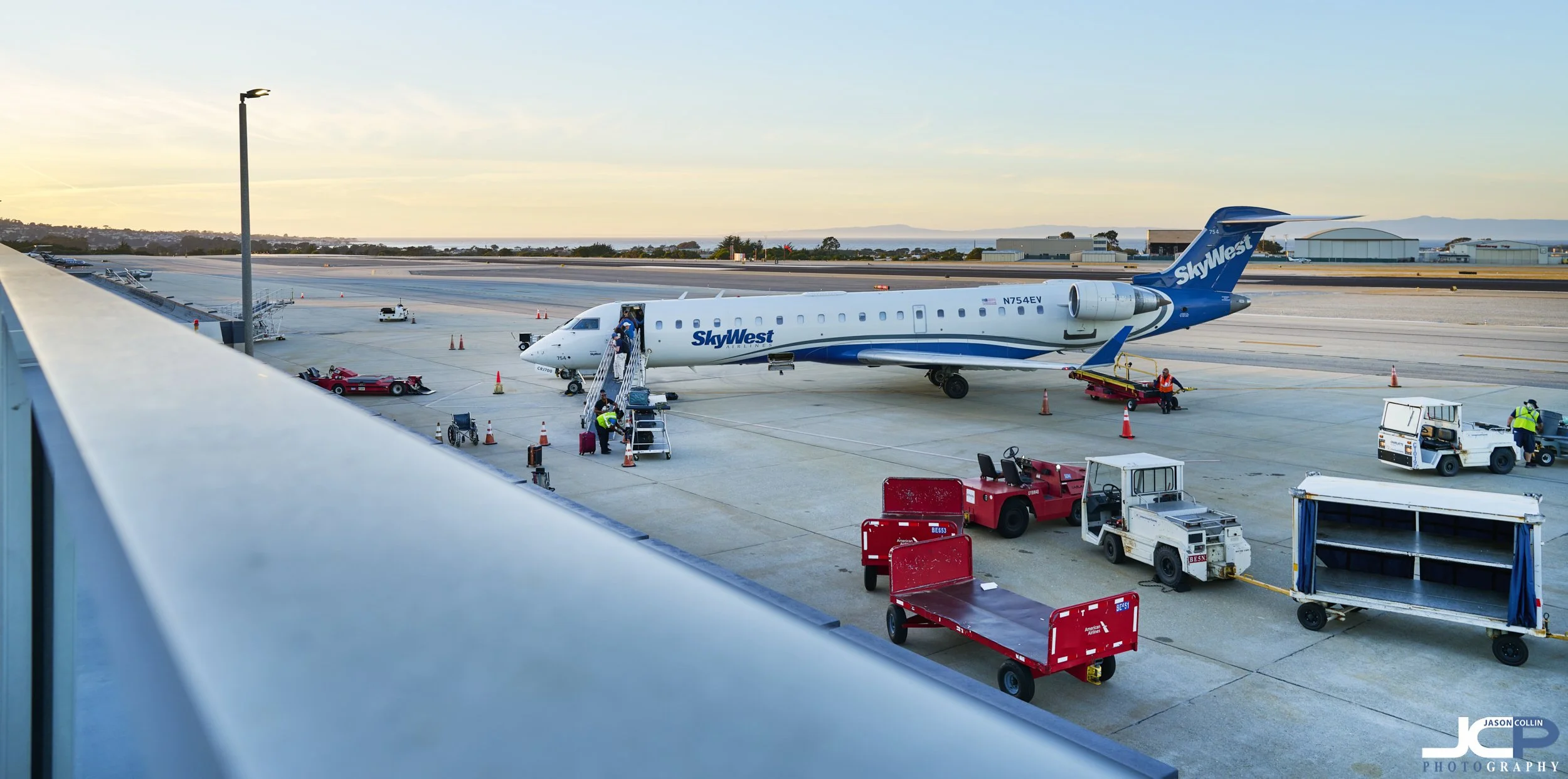 Commercial-style photograph of a SkyWest airplane at Monterey Regional Airport during twilight, with ground crew, equipment, and a leading line foreground composition overlooking the runway.