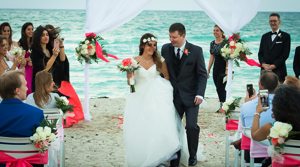 bride-groom-beach-wedding-ceremony-walking-aisle-happy-smiles-white-arch-flowers-pink-green-ribbons-chairs-sandy.jpg