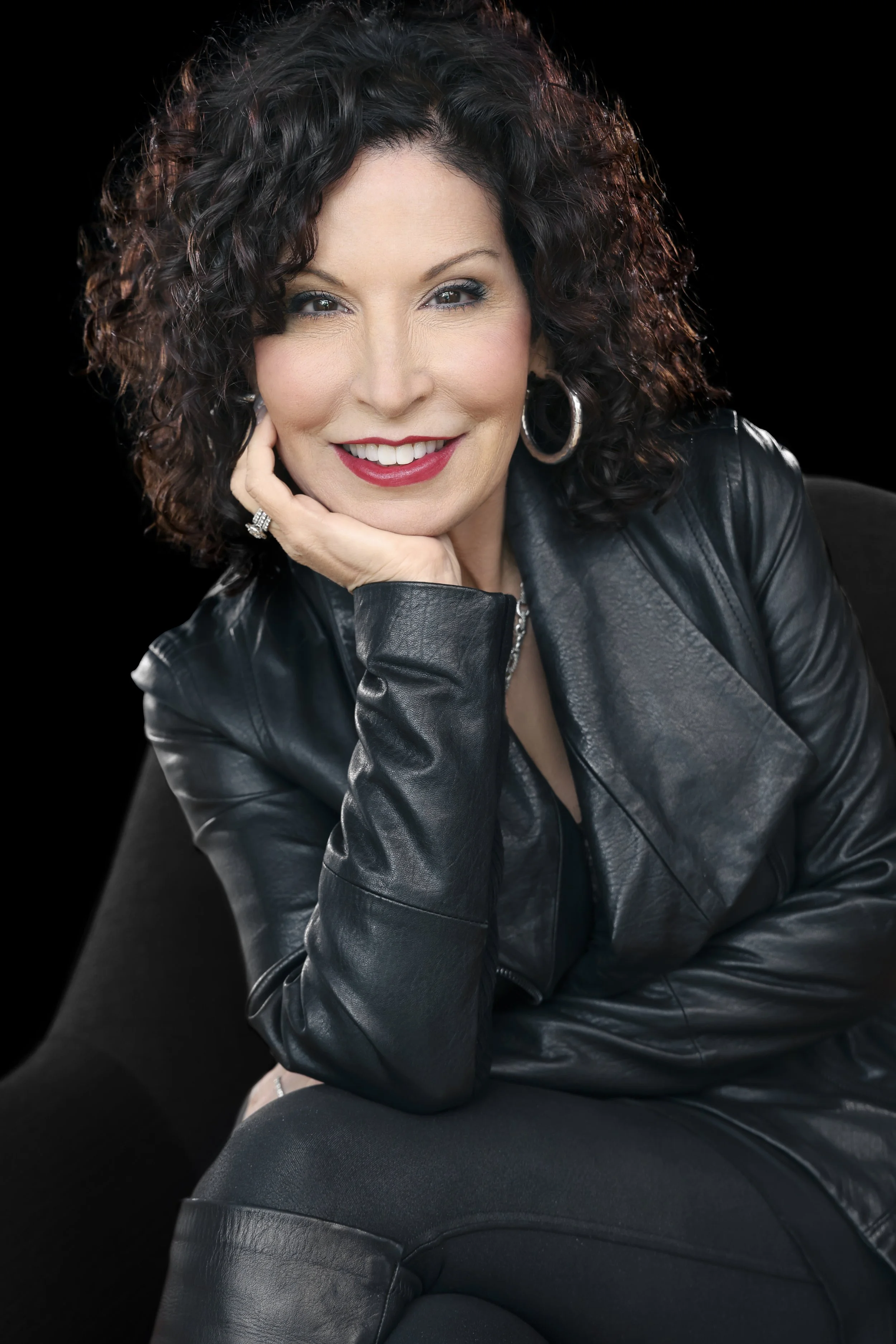 A woman with curly dark hair, wearing a leather jacket, smiling and resting her chin on her hand, sitting on a black chair against a black background.